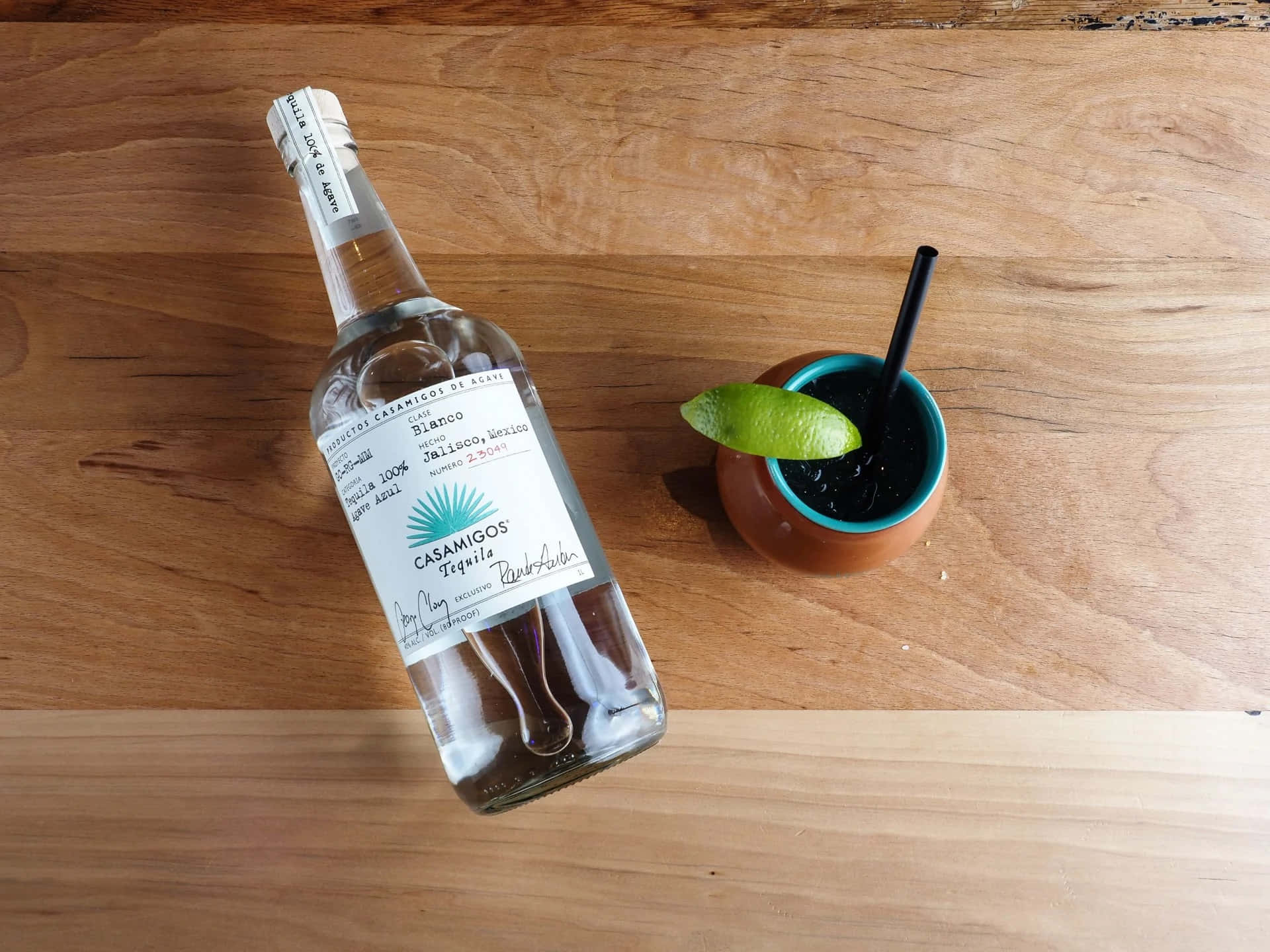 品味非凡：Casamigos 白色龍舌蘭酒馬提尼雞尾酒 壁紙 — 一瓶Casamigos Blanco龍舌蘭酒放在一個鮮藍色邊緣的陶杯旁邊，杯內盛載著深色雞尾酒，飾以青檸角，置於溫暖的木質表面上。