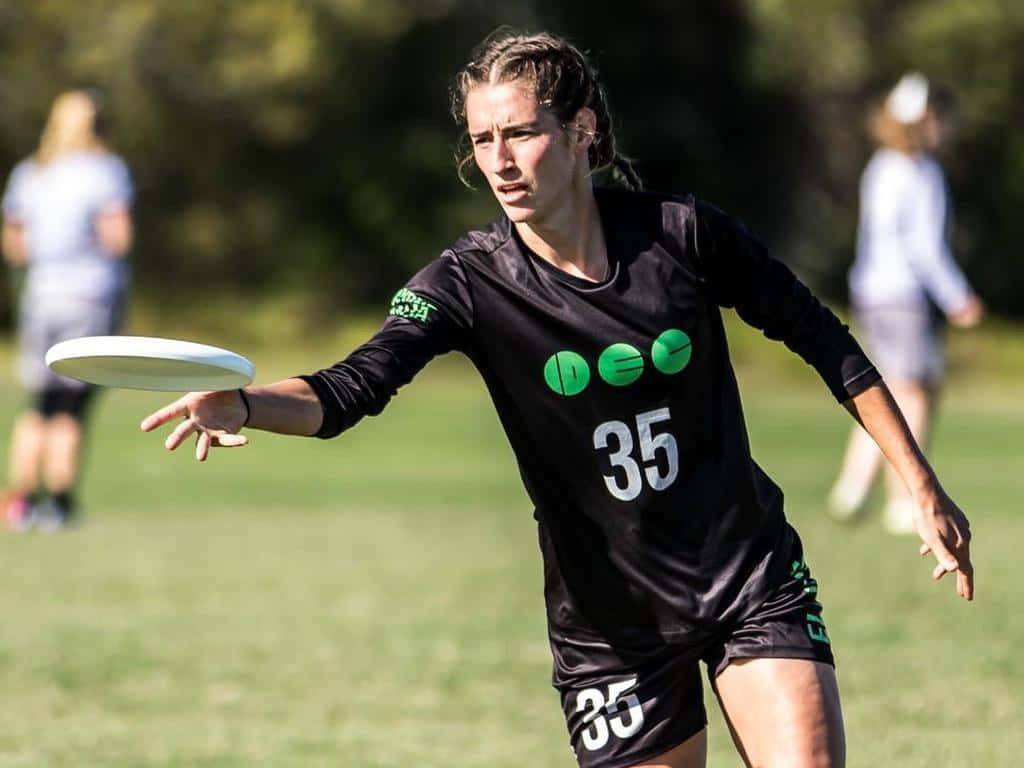 Cat Phillips 最佳 Ultimate Frisbee 背景 壁紙 — 一位專注的女運動員身穿黑色球衣，號碼35，將白色飛盤擲過陽光明媚的草地。