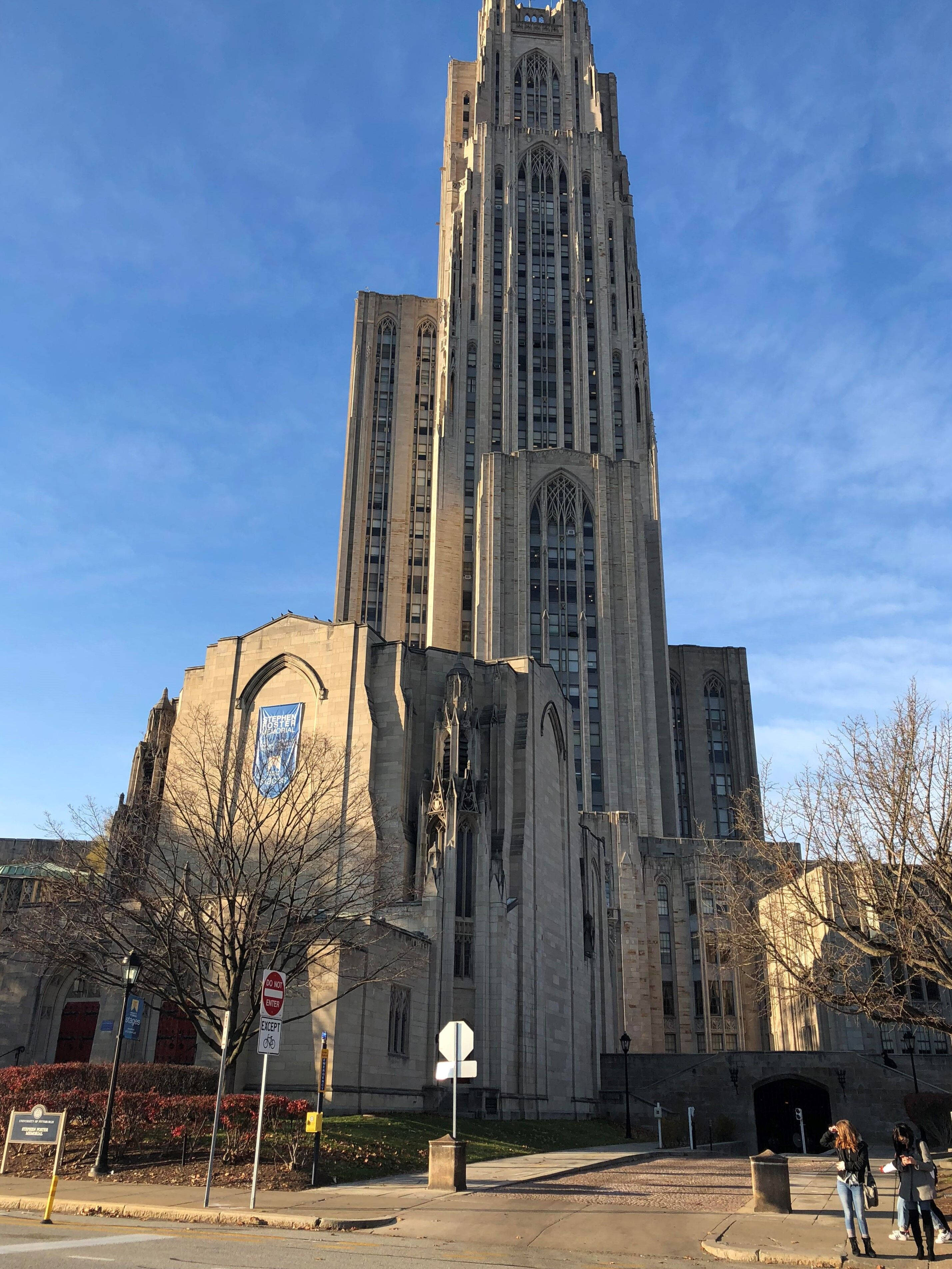 匹茲堡大學 Cathedral of Learning 建築壯觀景觀 壁紙 — 一座高聳的哥德式大學建築，擁有石頭外牆和高尖塔，在晴朗的藍天下，附近有光禿禿的樹木。