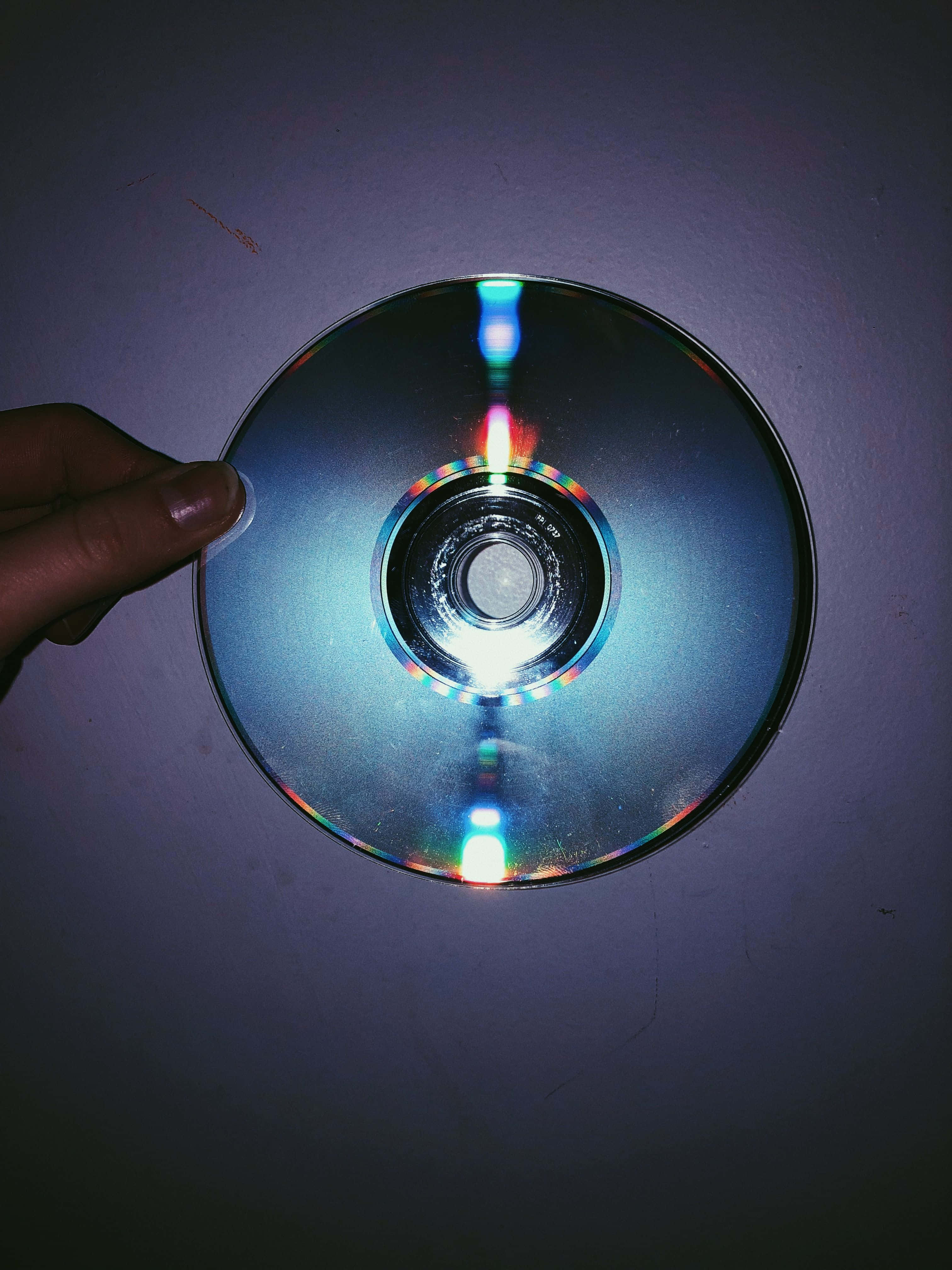 藍色龐克風格CD電影攝影 壁紙 — 一隻手拿著反光的CD靠在昏暗的紫色牆上，在情緒化的特寫中捕捉到色彩繽紛的光暈。