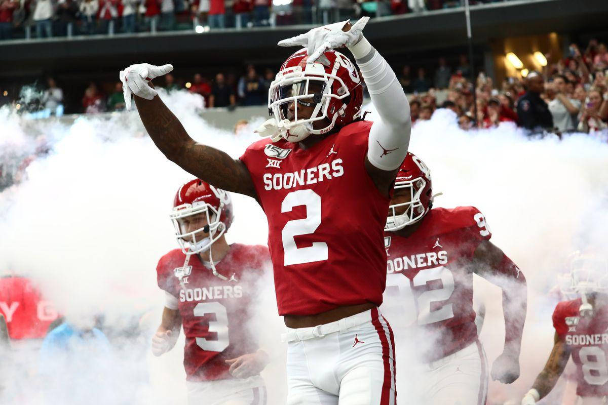 Ceedee Lamb 與隊友 Sooners 壁紙 — 一名穿著紅色Sooners裝備的足球員在球場上舉起雙臂慶祝，煙霧在他周圍翻騰。