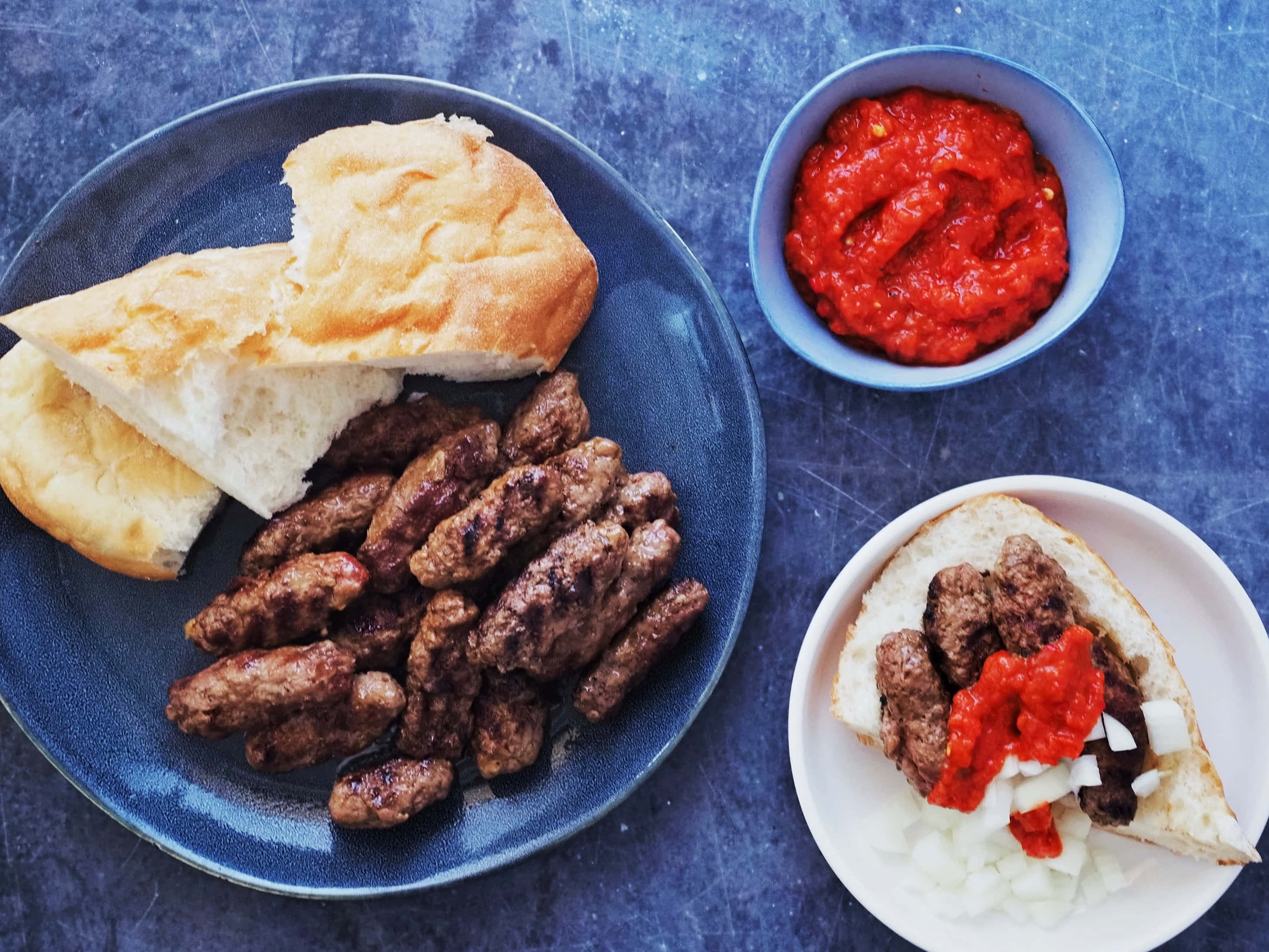 配溫熱皮塔麵包的傳統Ćevapi 壁紙 — 烤製肉腸配上新鮮麵包和鮮紅色醬汁，置於有紋理的藍色表面上。