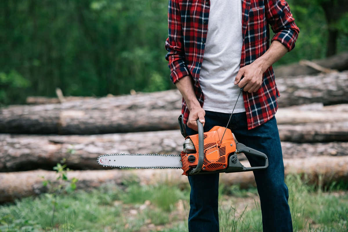 Chainsaw 在運作中 壁紙 — 一名身穿紅色格仔衫嘅男人喺茂密嘅綠色森林中，喺堆疊嘅圓木之間揸住一把橙色嘅鏈鋸。