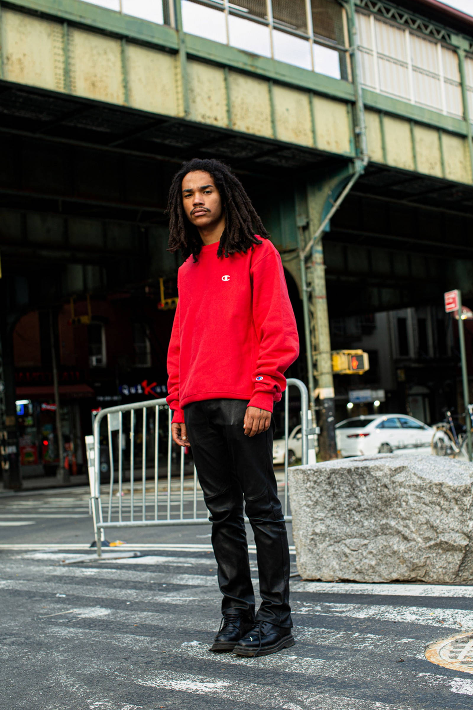 Champion 與 Luka Sabbat 聯名系列 壁紙 — 一位留著辮子的年輕男子自信地穿著紅色運動衫，站在有塗鴉和城市燈光的城市天橋下。