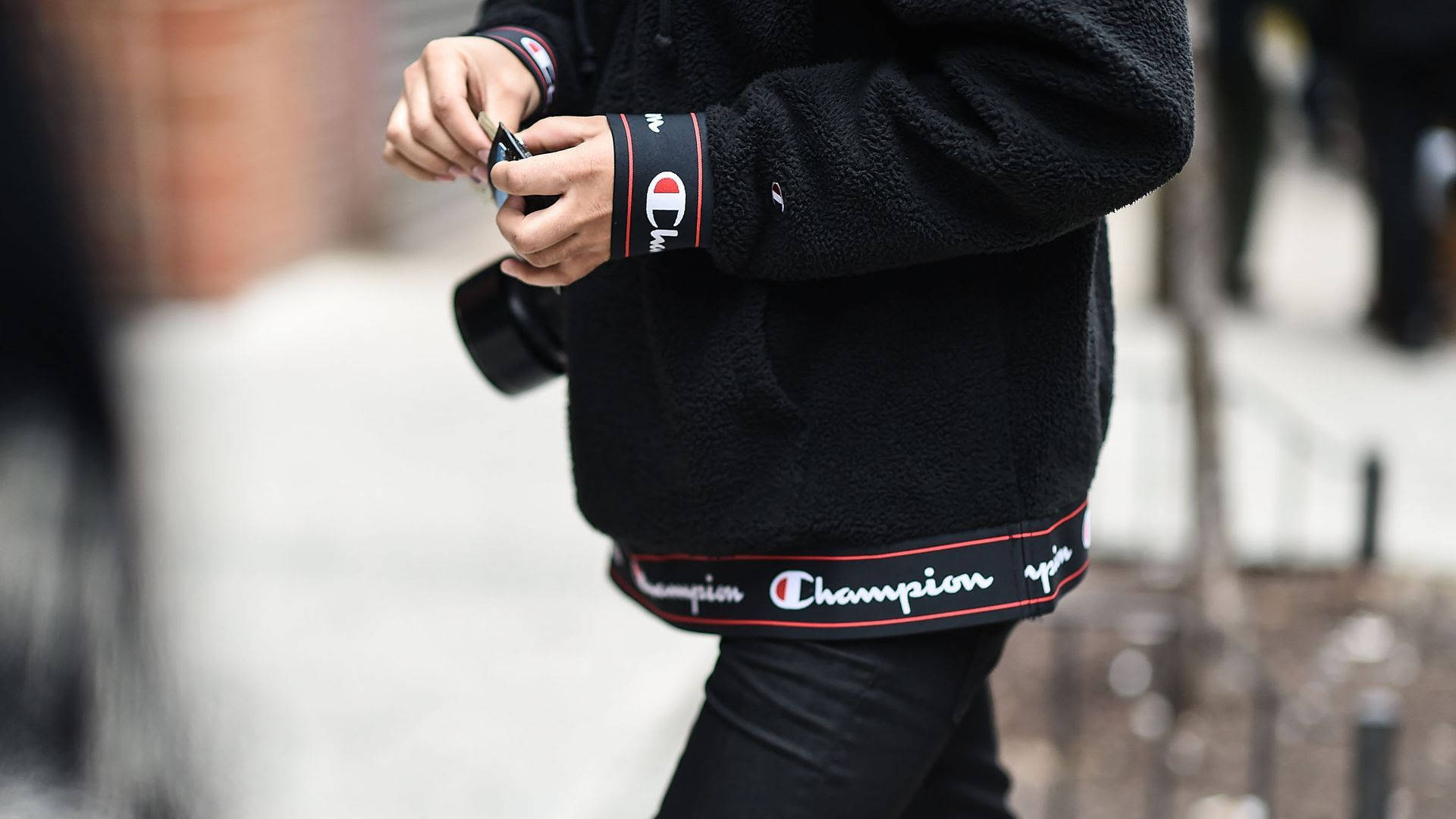Champion夾克內裡 壁紙 — 近距離拍攝一名身穿黑色Champion抓毛絨外套的人，在行人路上調整相機帶。