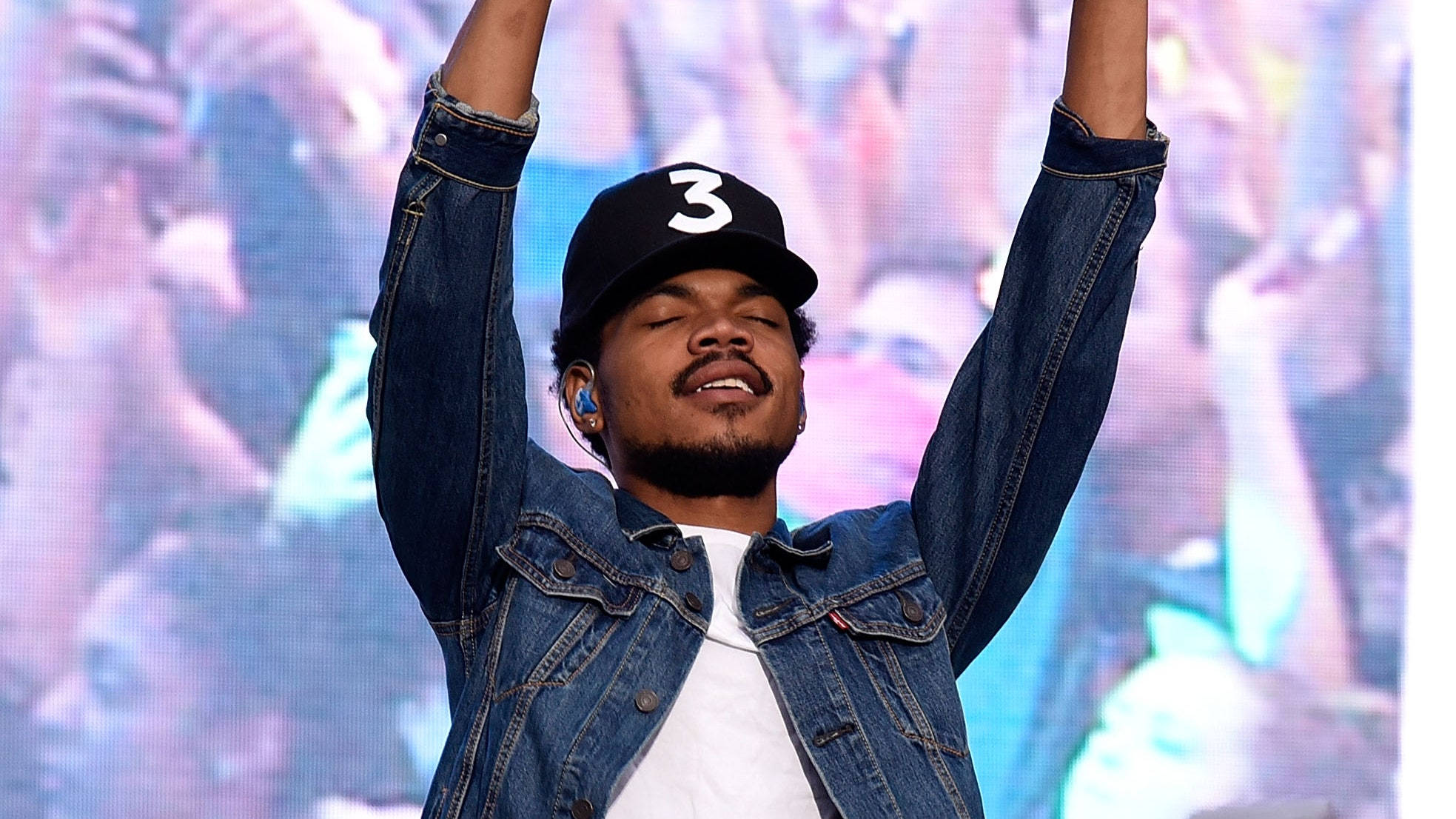 Chance The Rapper 全神投入 壁紙 — 一位身穿牛仔外套和帽子的男性表演者在舞台上，在閃耀的人群前勝利地舉起雙臂。