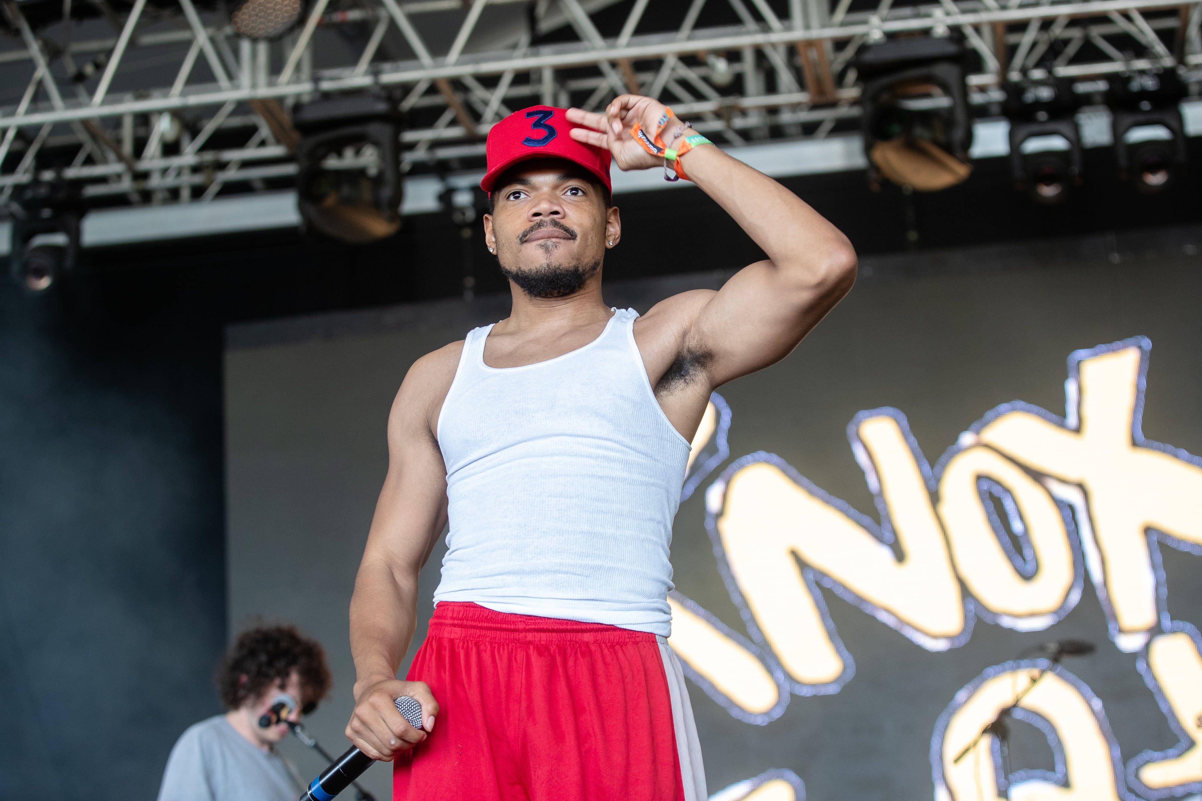 Chance The Rapper 展現肌肉 壁紙 — 一位戴紅色帽、身穿白色背心的男表演者在舞台上，在明亮燈光及發光背景下手持咪高峰。