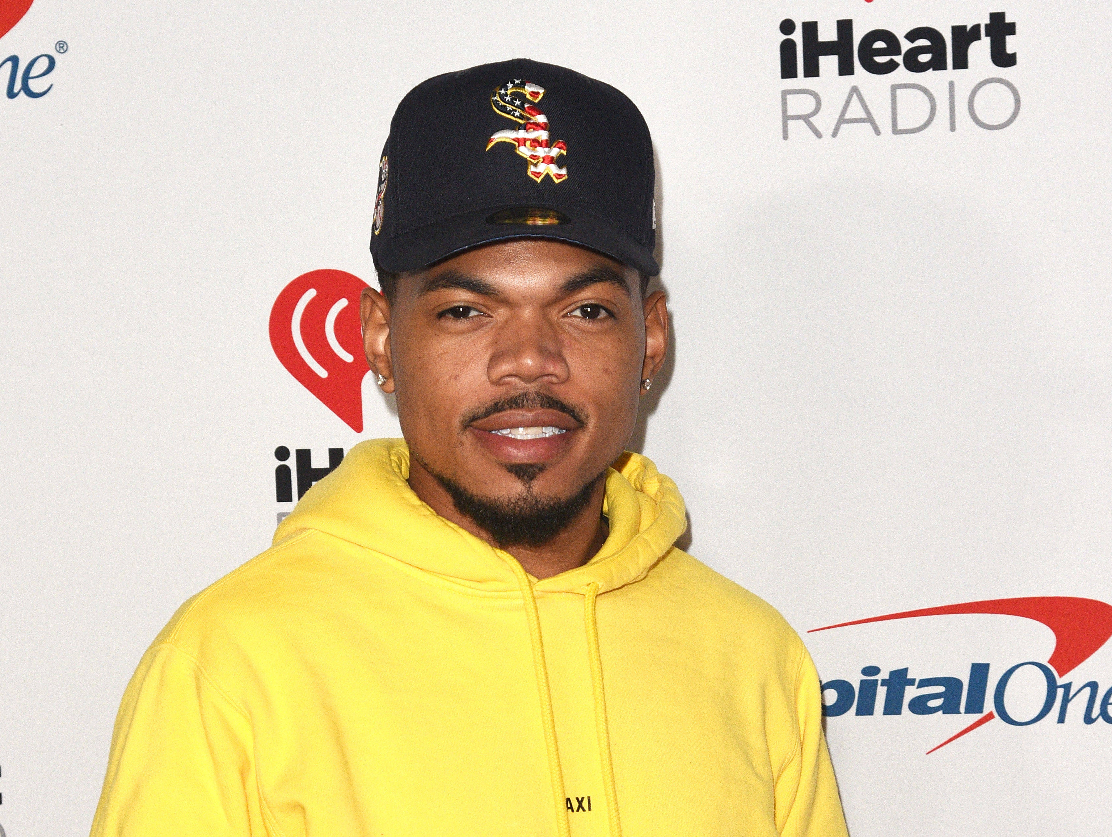 Chance The Rapper 黃色連帽衫 壁紙 — 一名身穿亮黃色連帽衫、戴黑色帽子的男子在品牌活動背景前對著鏡頭微笑。