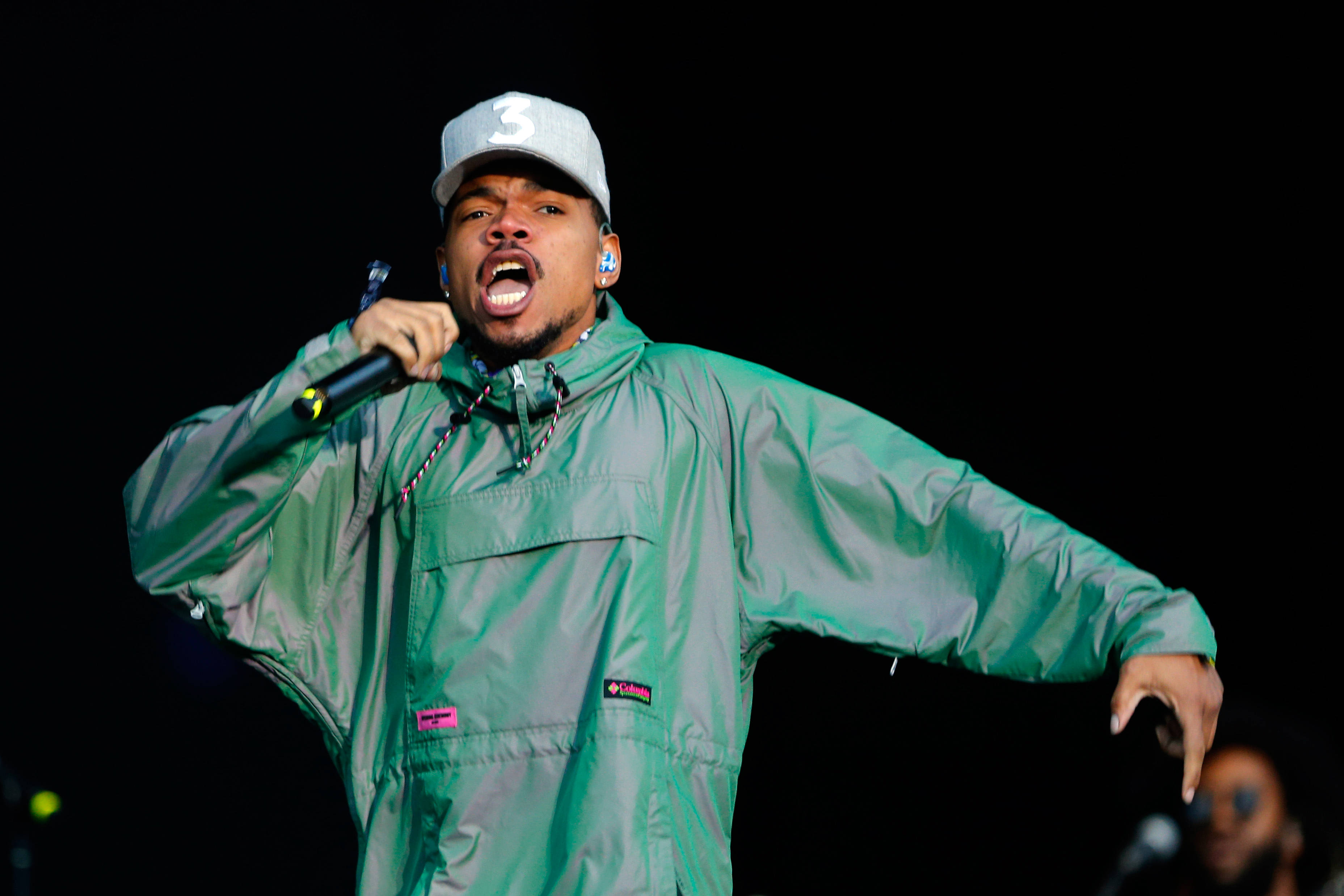 Chance the Rapper綠色連身褲 壁紙 — 一位身穿綠色外套和白色帽子的男性饒舌歌手，在黑暗的舞台背景下熱情地對著麥克風表演。