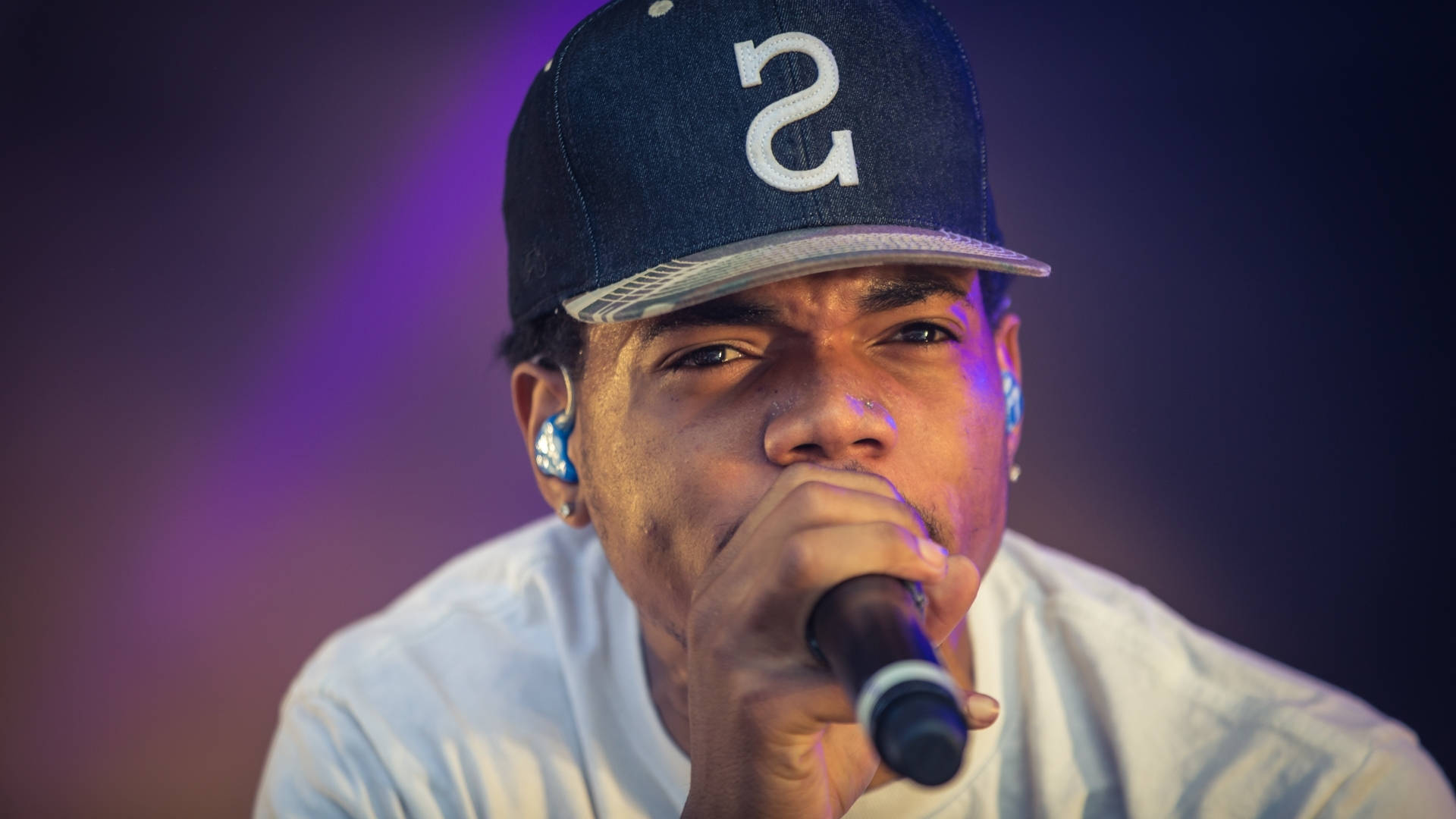 Chance The Rapper 現場演出 壁紙 — 一位專注的饒舌歌手戴著帶有白色「S」標誌的海軍藍帽子，在充滿活力的紫色舞台燈光下將麥克風靠近嘴邊。