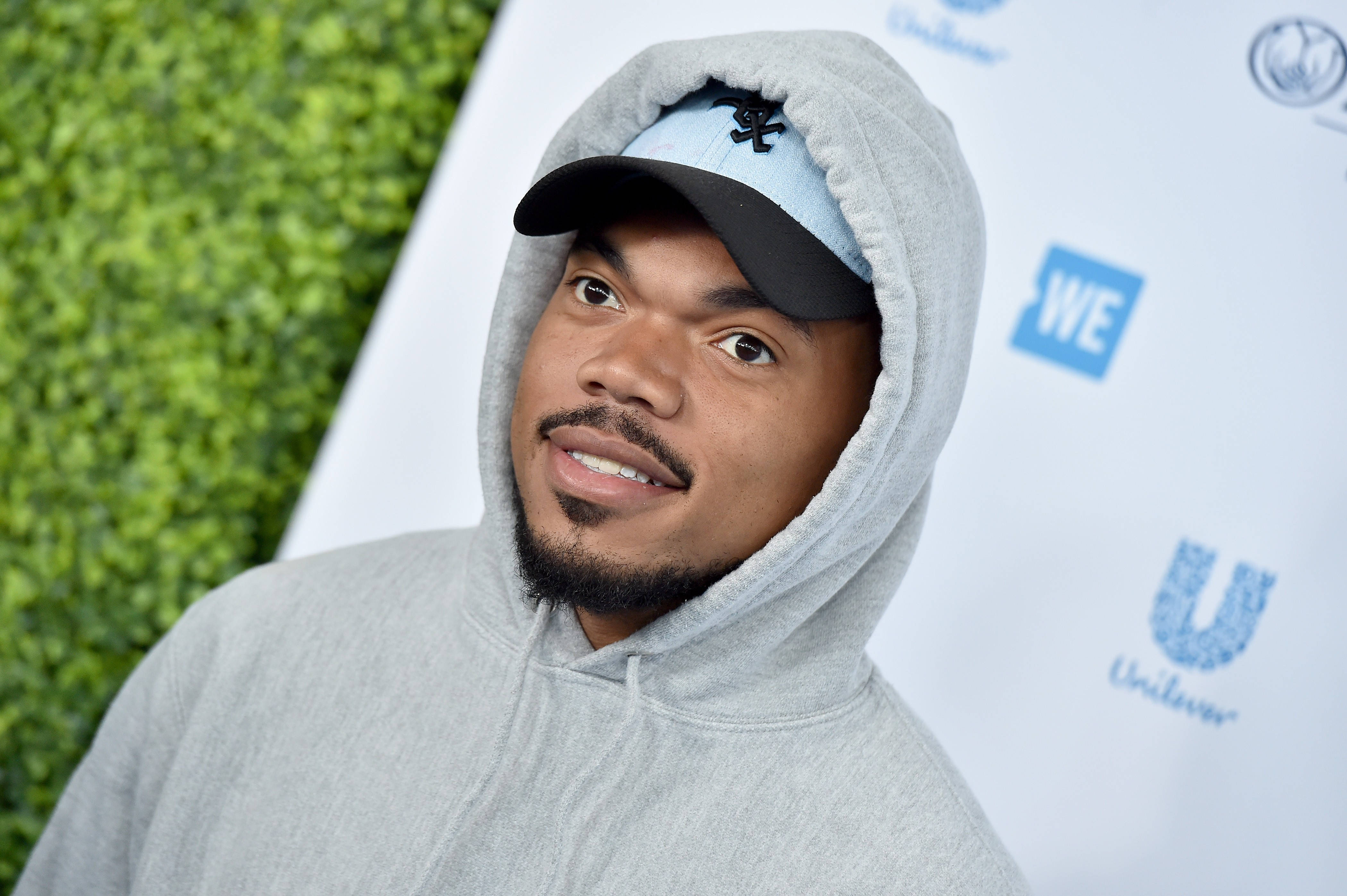 Chance The Rapper 灰色毛衣 壁紙 — 一名穿著灰色連帽衫和帽子的男子在綠色樹籬和品牌背景前對著鏡頭微笑。