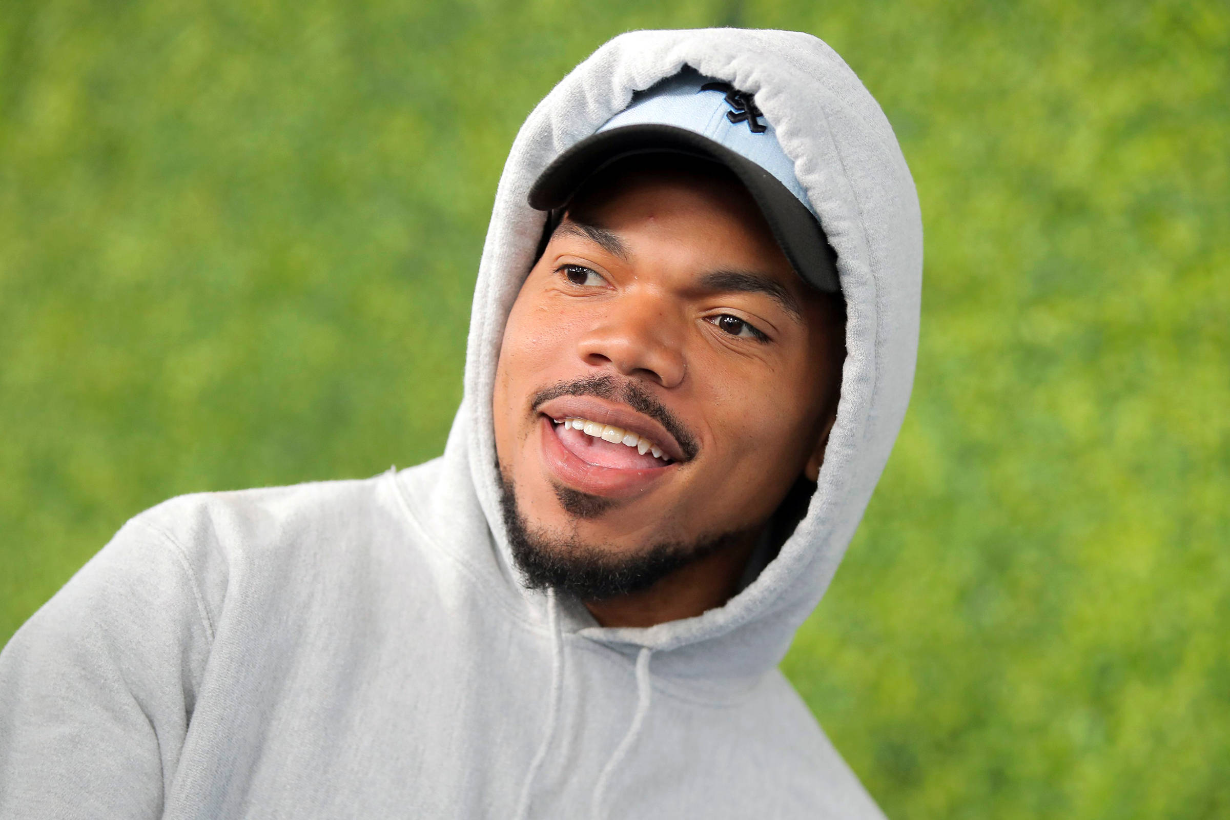 Chance the Rapper綠色草地 壁紙 — 一個微笑的男人，身穿灰色連帽衫和帽子，背景是充滿活力的綠色模糊背景。