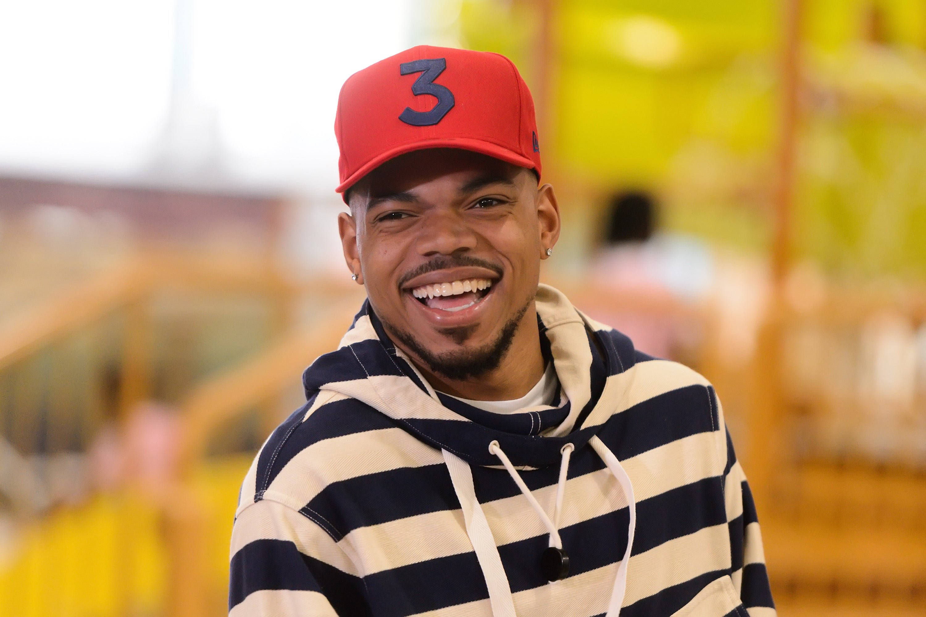 Chance the Rapper條紋連帽衫 壁紙 — 一位微笑的男子戴著紅色帽子，上面有黑色數字三，身穿條紋連帽衫，在模糊多彩的背景前開懷大笑。
