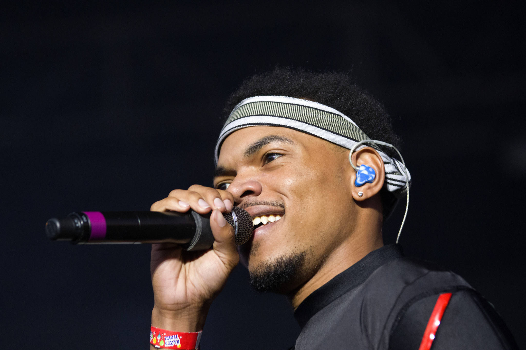 Chance The Rapper 白色頭帶 壁紙 — 一位微笑的男性表演者在舞台燈光下對著咪高峰唱歌，背景昏暗。