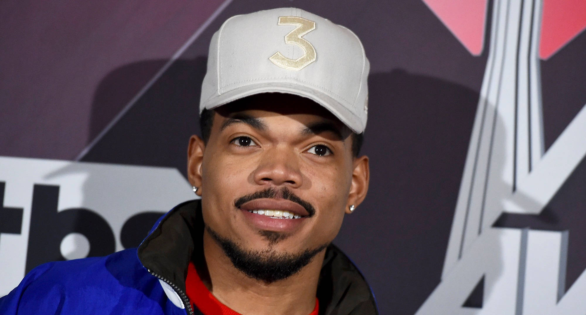 Chance The Rapper 微笑 壁紙 — 一位留著山羊鬍、戴著金色三號帽子的男士在品牌背景前對著鏡頭微笑。