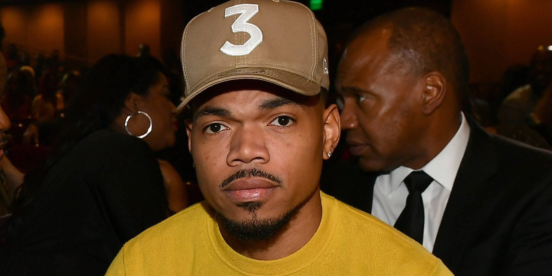 Chance The Rapper 橘黃色上衣 壁紙 — 一名身穿黃色襯衫、戴著米色帽子、帽上有白色數字三的男子，在昏暗的室內環境中直視鏡頭。