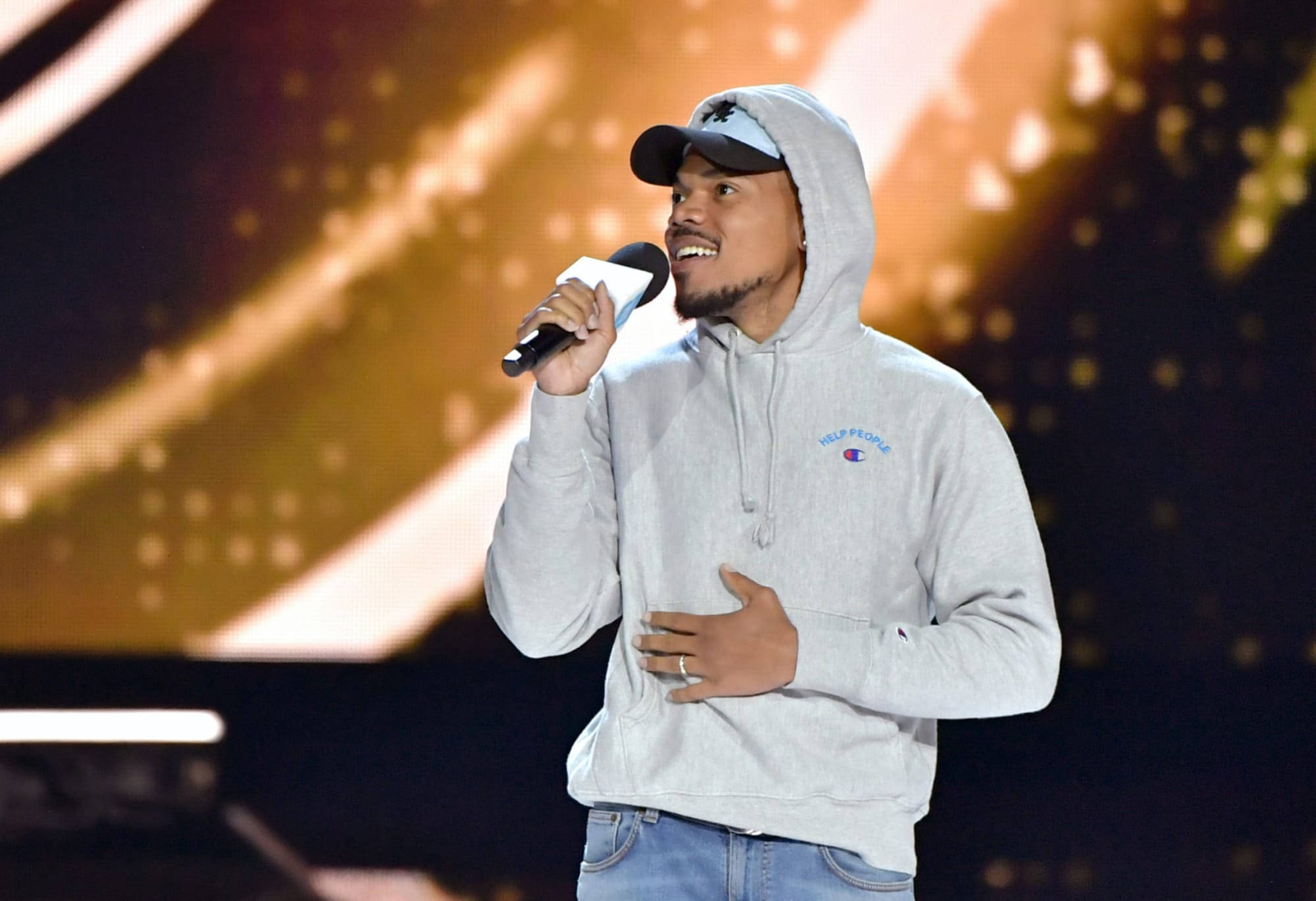 Chance the Rapper幫助他人 壁紙 — 一位微笑的男子身穿灰色連帽衫和帽子，在金色光束和散景效果的舞台上對著麥克風唱歌。