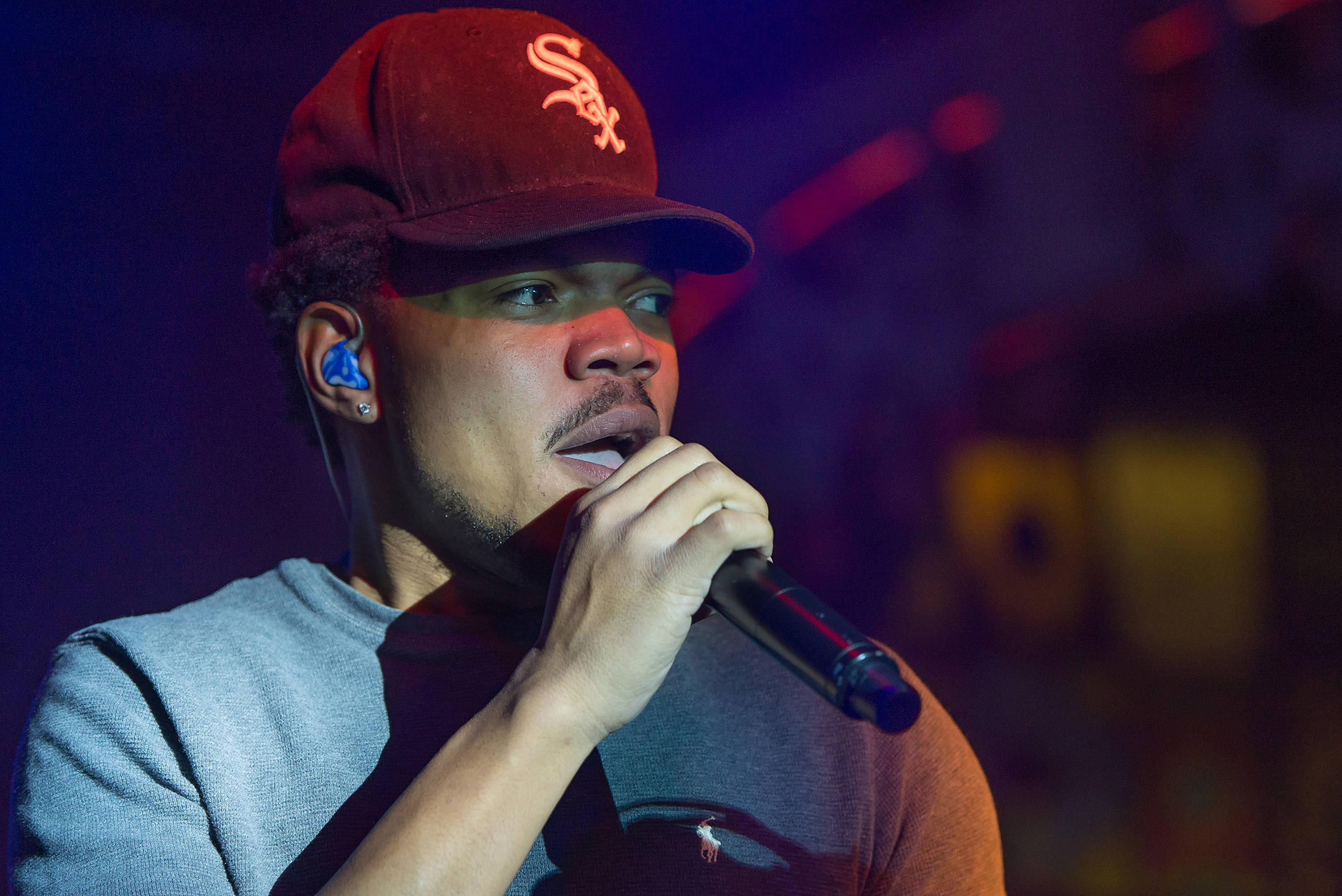 Chance The Rapper 性感帽款 壁紙 — 一位戴著紅色棒球帽的男說唱歌手在充滿活力的舞台燈光下對著麥克風唱歌。