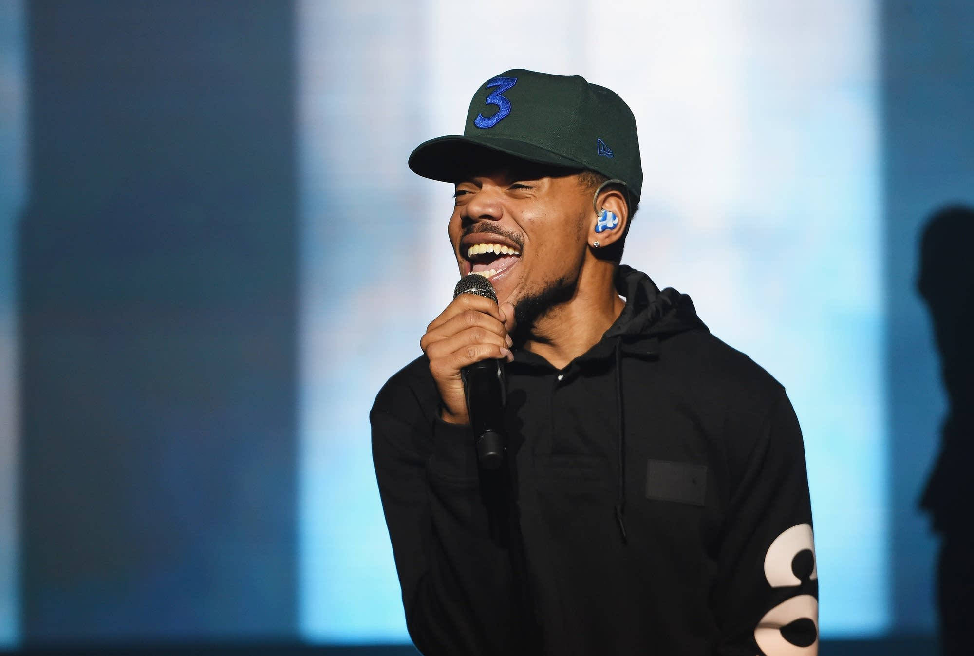 活力四射的Chance the Rapper 壁紙 — 一位微笑的表演者戴着绿色帽子和黑色连帽衫，在明亮的舞台灯光下对着麦克风唱歌。
