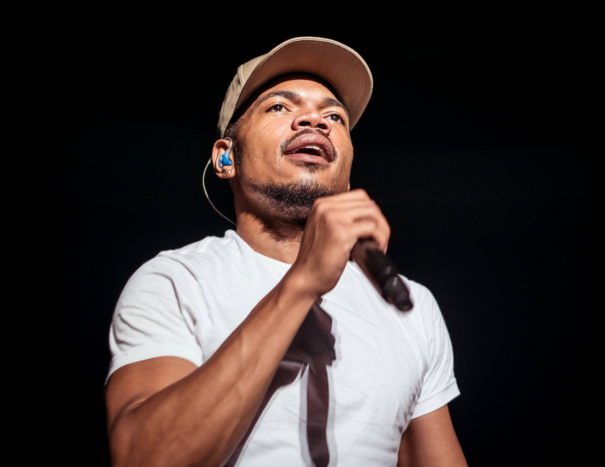 Chance the Rapper演出 壁紙 — 一位身穿白色T恤和米色帽子的男歌手，在黑色背景前對著麥克風充滿激情地演唱。