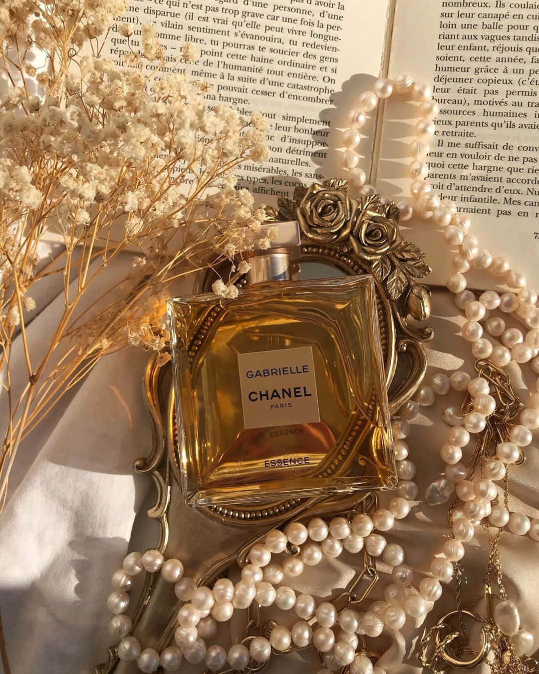  壁紙 — Chanel Gabrielle Essence 香水瓶在華麗的鏡子上閃耀，周圍環繞著珍珠、乾花和打開的書頁，沐浴在溫暖的陽光下。
