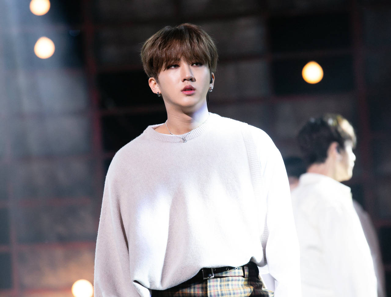 Changbin K-Pop 偶像 壁紙 — 一位身穿白色毛衣和格子褲的年輕男子自信地站在燈光昏暗的舞台上，身後溫暖的聚光燈閃耀著。