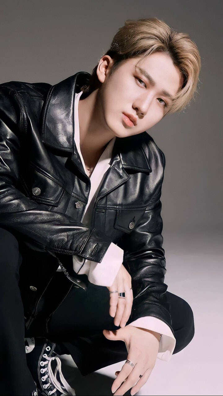 Changbin 韓國名人 壁紙 — 一位年輕男子，留著時尚的金色短髮，身穿黑色皮褸內搭白襯衫，蹲在灰色中性背景前。