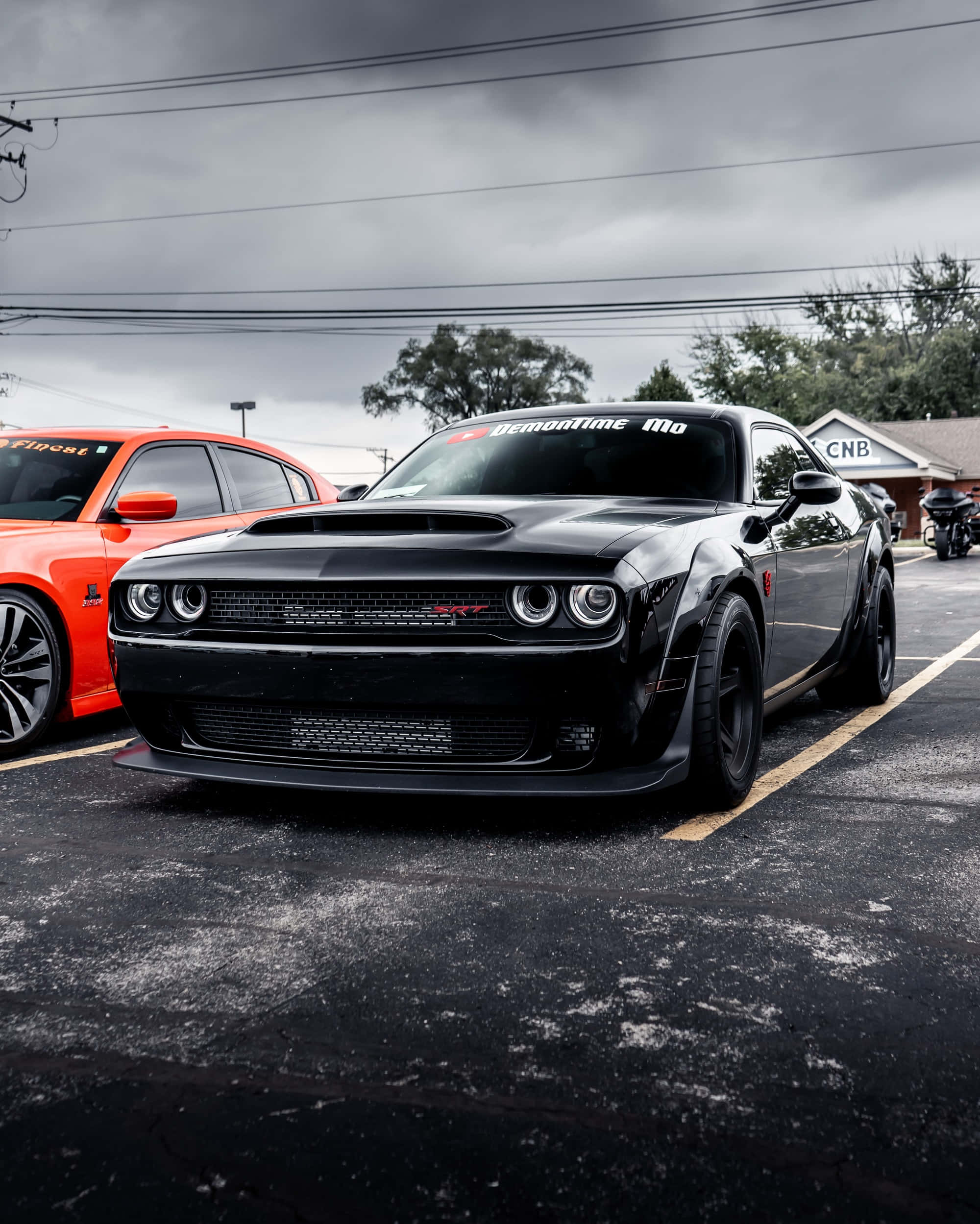 經典道奇挑戰者與 Charger 壁紙 — 一輛黑色Dodge Challenger Hellcat帶有'DemonTime Mo'貼紙，停泊在一個橙色汽車旁邊的車位，天空多雲。