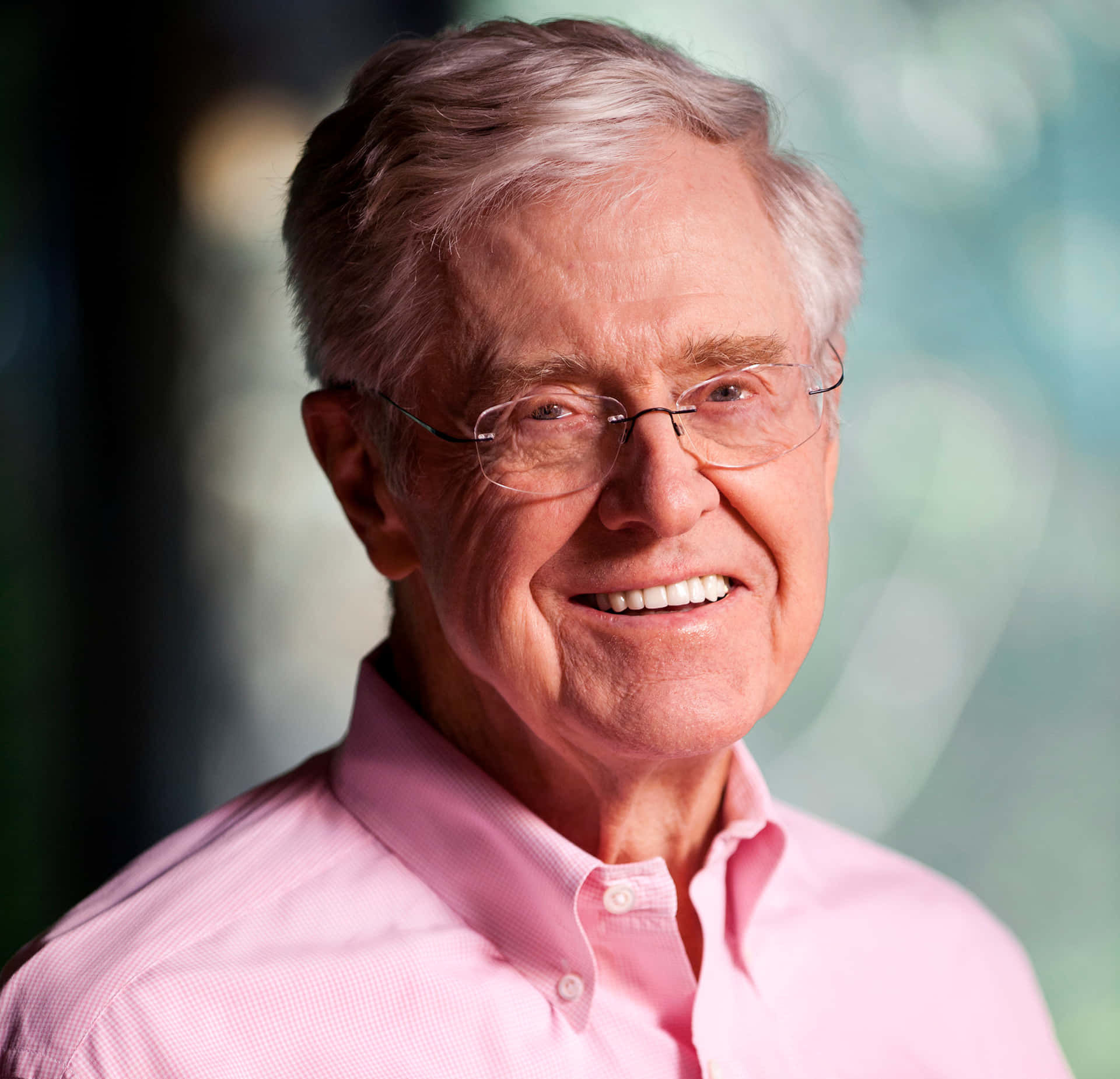 Charles G. Koch 穿粉紅襯衫微笑 壁紙 — 一位戴眼鏡、灰白頭髮的開朗長者，穿著粉紅色鈕扣襯衫，在柔和模糊的背景前溫暖地微笑。