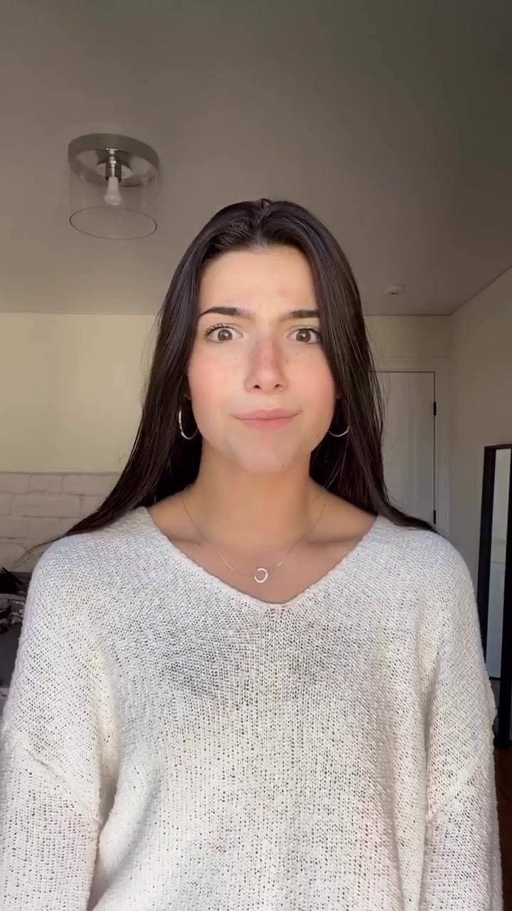 Charli Damelio 可愛表情 壁紙 — 一位長黑髮戴圈耳環的年輕女性，在室內柔和自然光下穿著質感白色毛衣。
