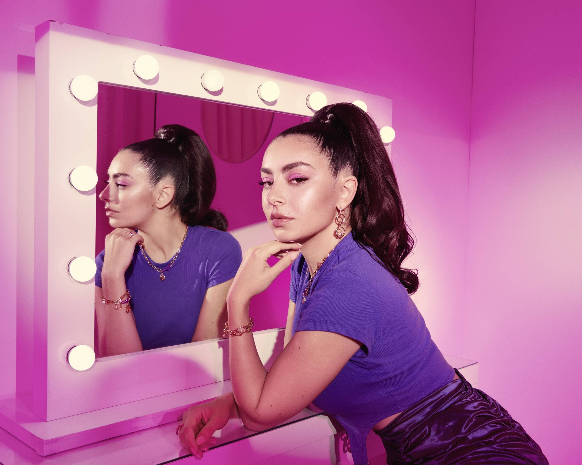 Charli XCX Pandora ME 首飾廣告 壁紙 — 一位身穿紫色服裝的女性，若有所思地凝視著自己在粉紅色燈光、帶有發光燈泡的梳妝鏡中的倒影。