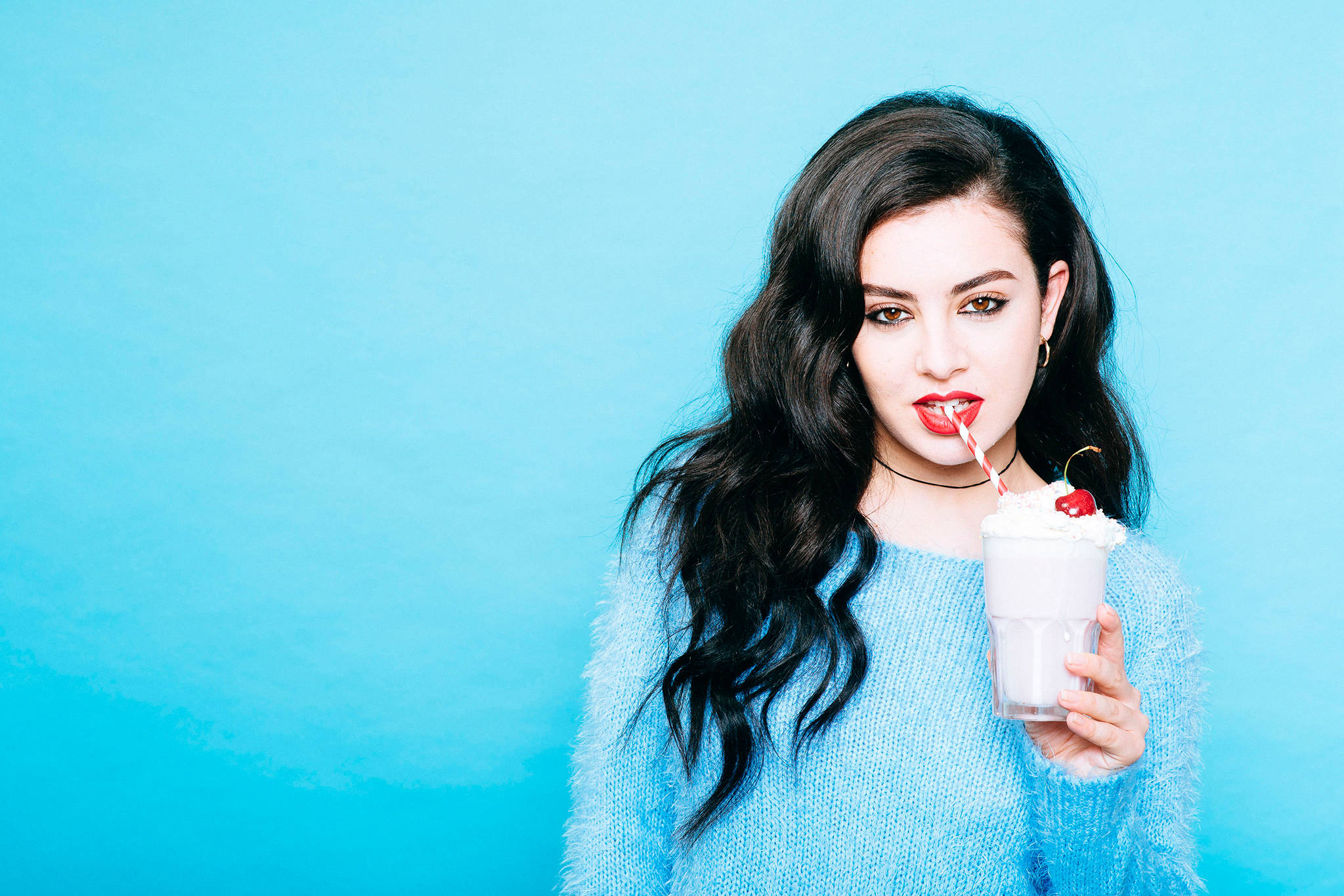 Charli XCX 藍色系美學 壁紙 — 一位擁有深色波浪長髮的女性，在純藍色背景前啜飲一杯 topped with 忌廉和櫻桃的奶昔。