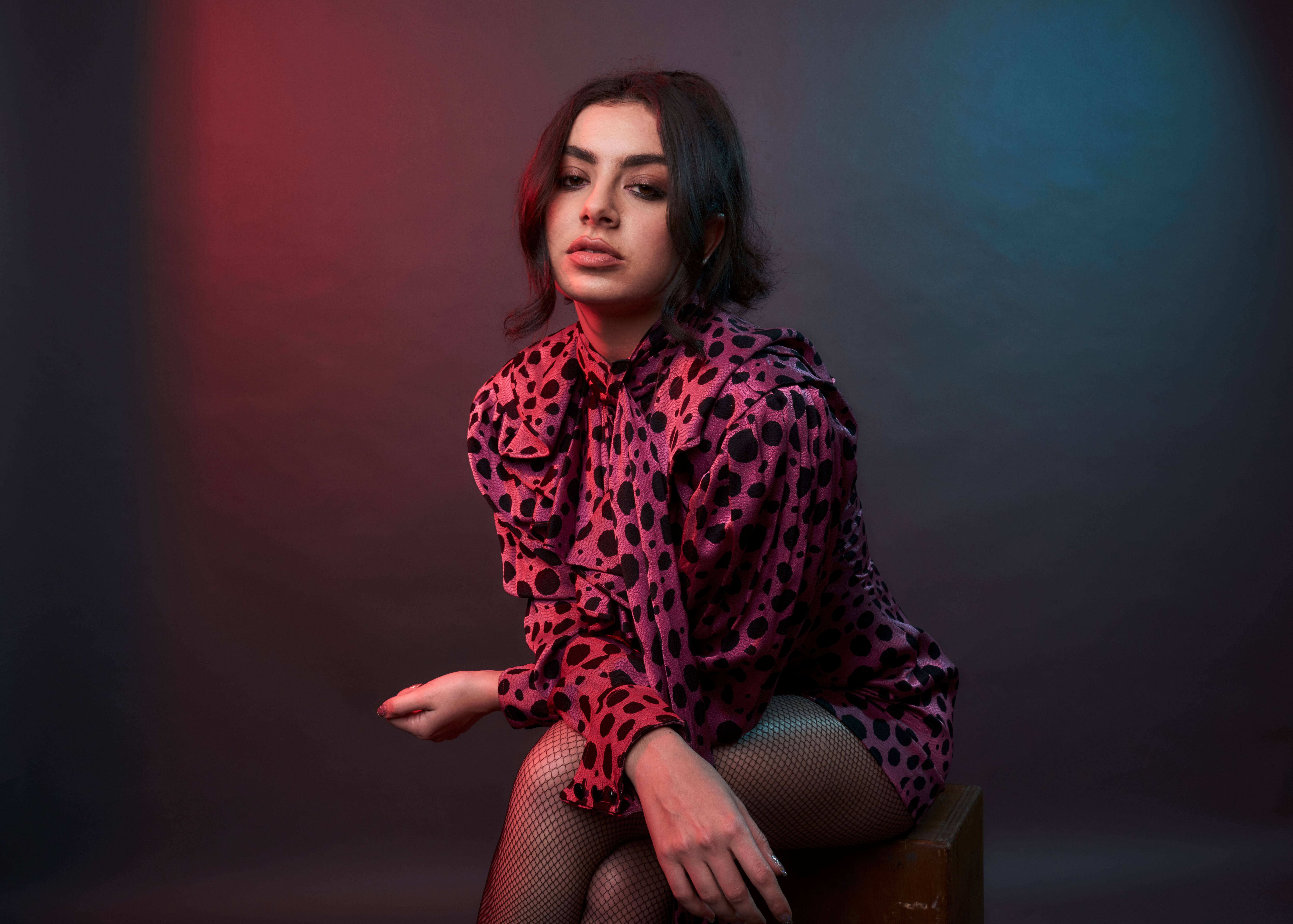 Charli XCX 豹紋圖案 壁紙 — 一位身穿粉紅色豹紋襯衫的女性，在充滿氛圍的紅藍燈光背景下優雅地坐著。
