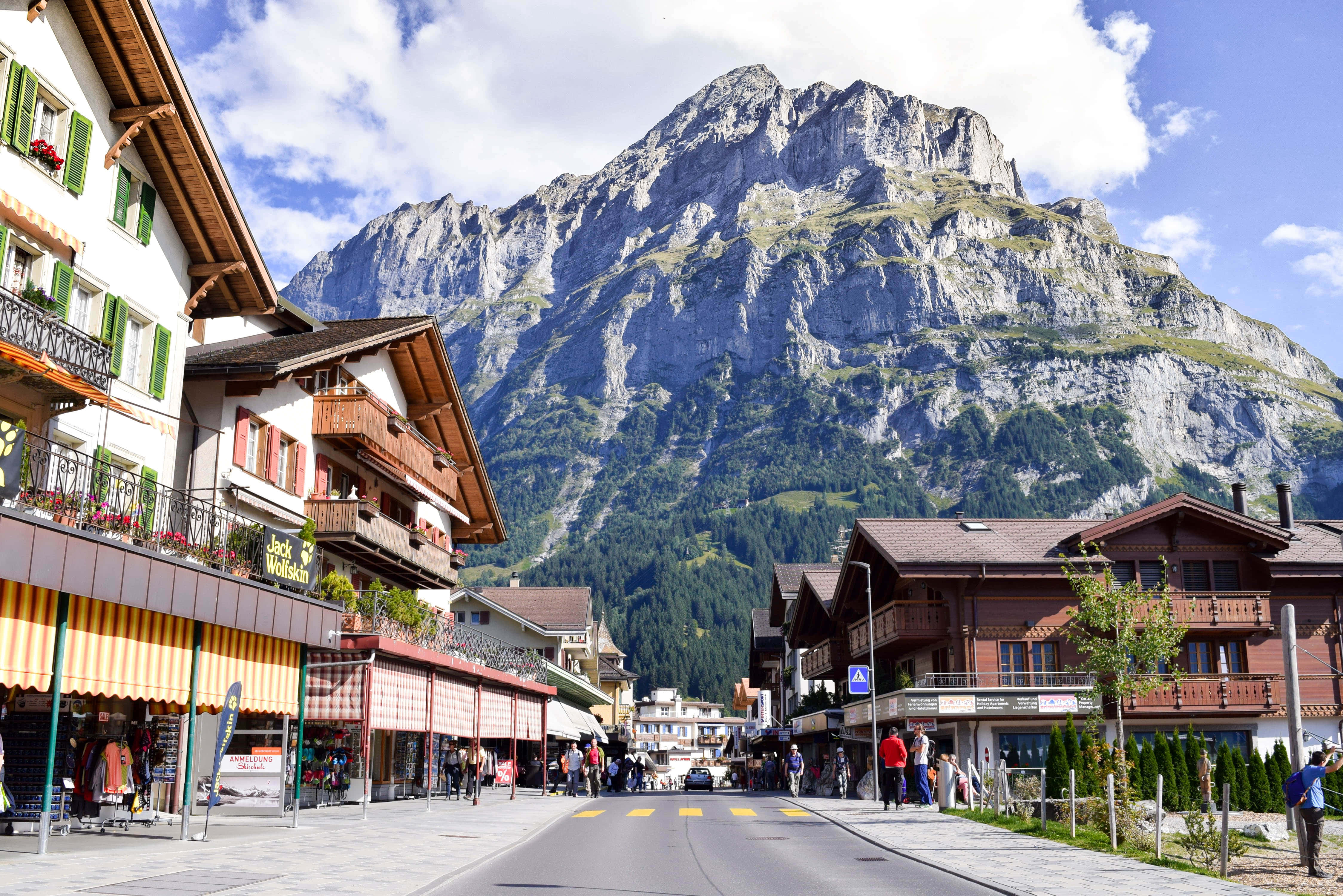 Switzerland 桌布 壁紙 — 一條迷人的山村街道，有傳統木質小屋和嶂峠的岩石山峰，上空是漂亮的藍天。