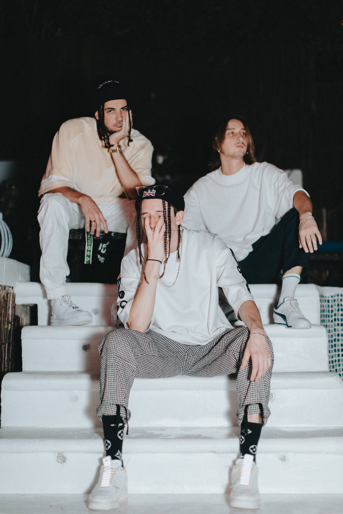 Chase Atlantic 將熱情注入音樂 壁紙 — 三位身穿白色服裝的時尚年輕男士在夜間燈光下，以深色背景為襯，隨意地坐在白色階梯上擺姿勢。