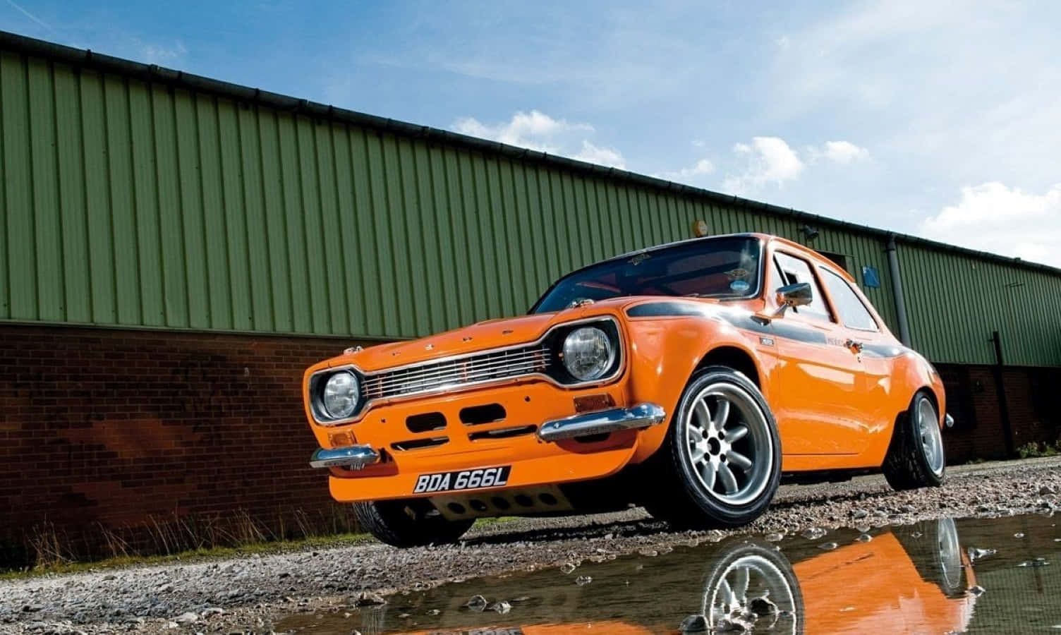 Ford Escort 桌布 壁紙 — 一輛充滿活力的橙色經典福特Escort拉力賽車在藍天下閃閃發光，其倒影在綠色金屬建築旁的水坑中閃爍。