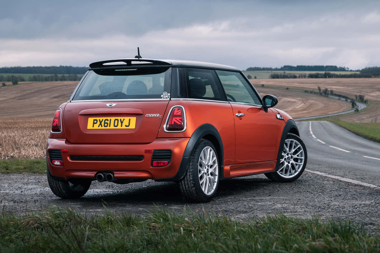 Mini Cooper S 桌布 壁紙 — 一輛充滿活力的橙色Mini Cooper S停泊在碎石路邊，背景是蜿蜒的鄉村道路和陰天。