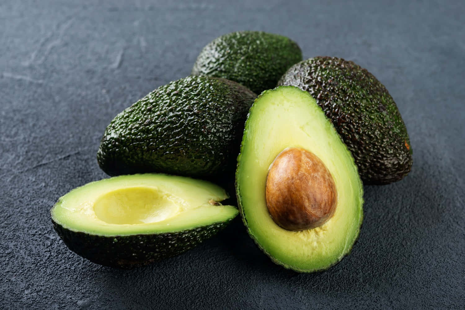 Avocado 桌布 壁紙 — 成熟的牛油果，其中一個切半露出奶油狀綠色果肉和棕色果核，放在深色紋理石面上。
