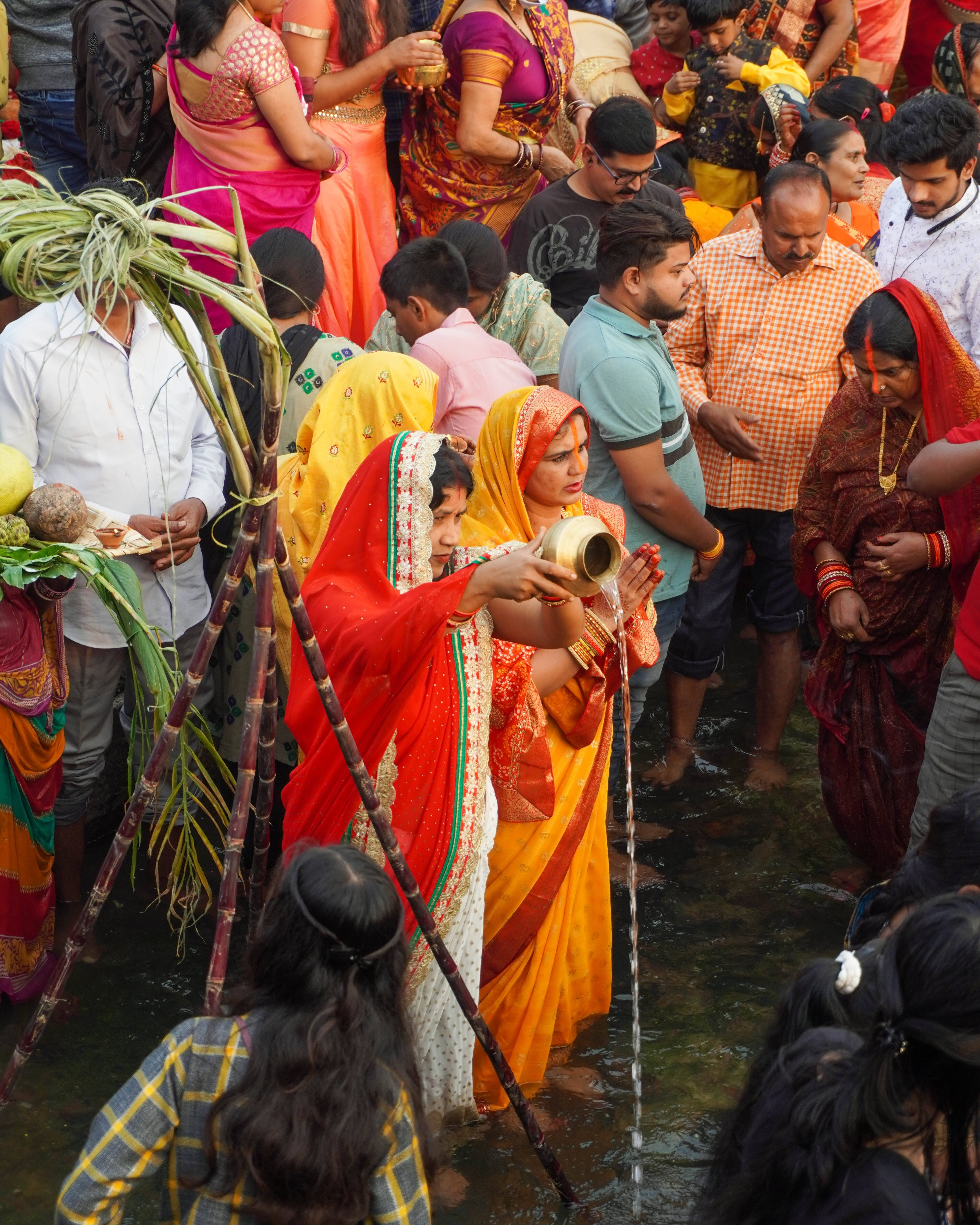 腳浸水中的人群 Chhath Puja 壁紙 — 一名身穿鮮豔橙色和紅色紗麗的女子，在擁擠的文化儀式中，將黃銅壺中的水倒入河中。