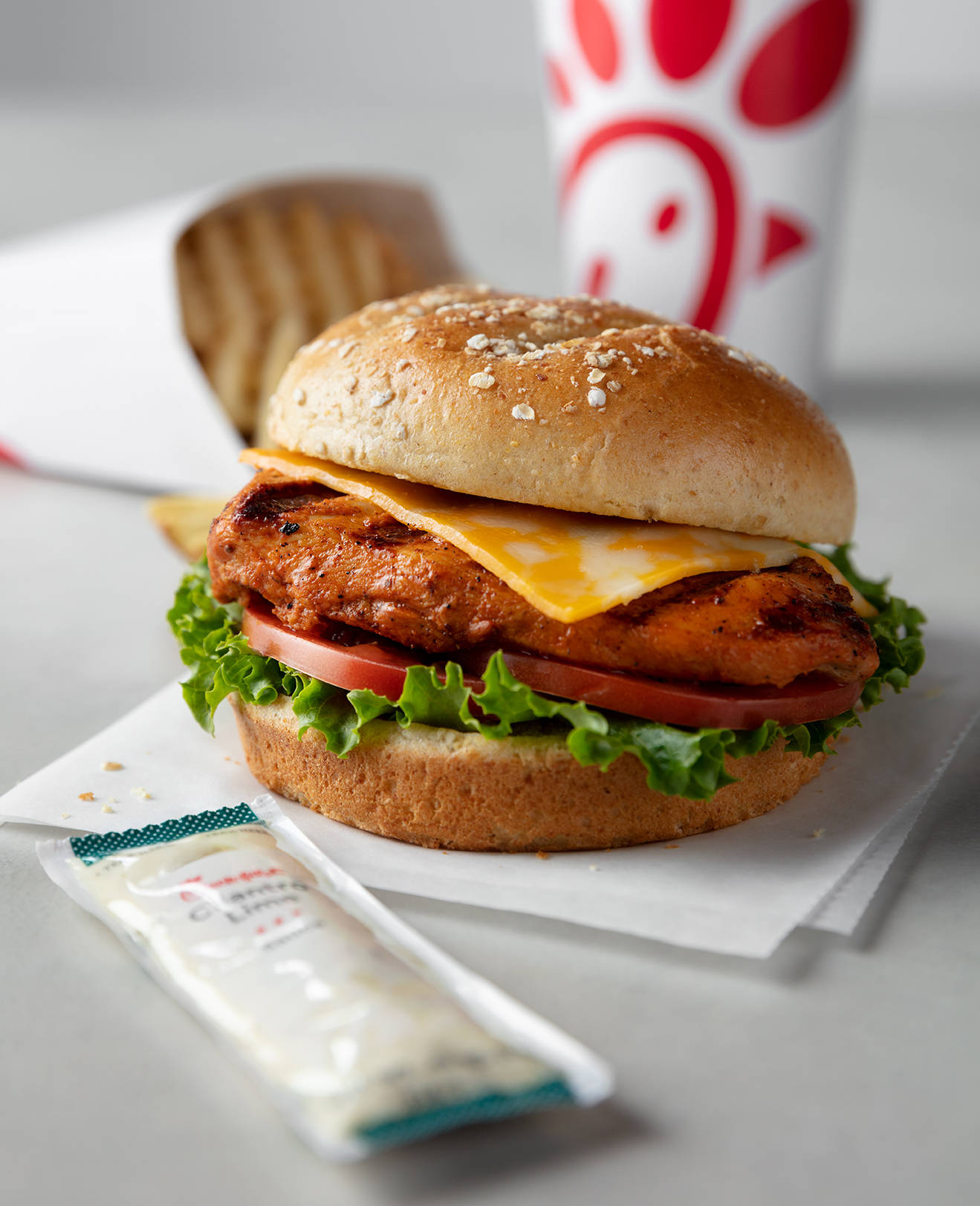 Chick Fil A 烤雞三明治 壁紙 — 一個脆皮雞肉三文治的近距離特寫，配有融化芝士、番茄和生菜，放在芝麻麵包上，旁邊有一個Chick-fil-A杯子和醬料包。