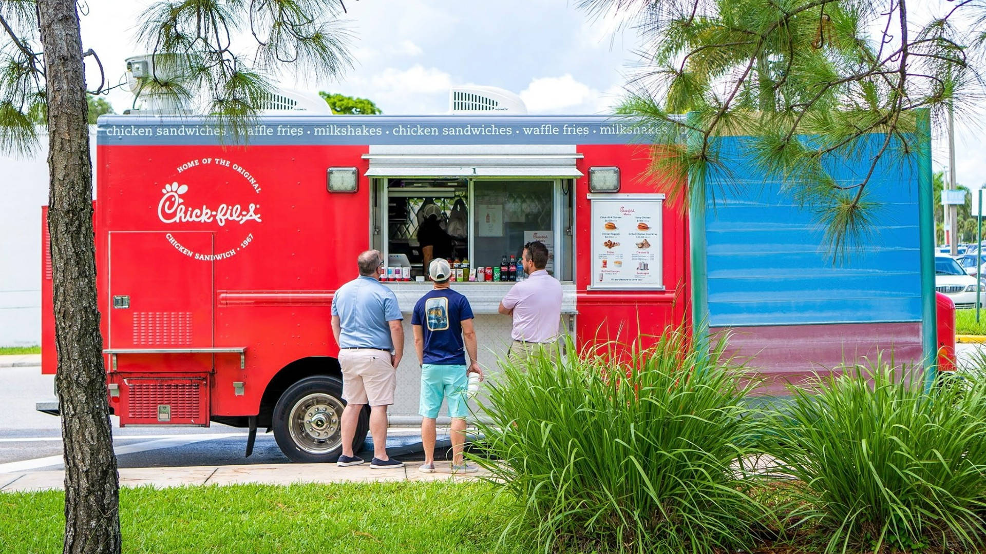 Chick Fil A 移動餐車 壁紙 — 三名男子站在鮮紅色的Chick-fil-A美食車前，在陽光明媚的天空下，被松樹和綠草環繞，正在點餐。