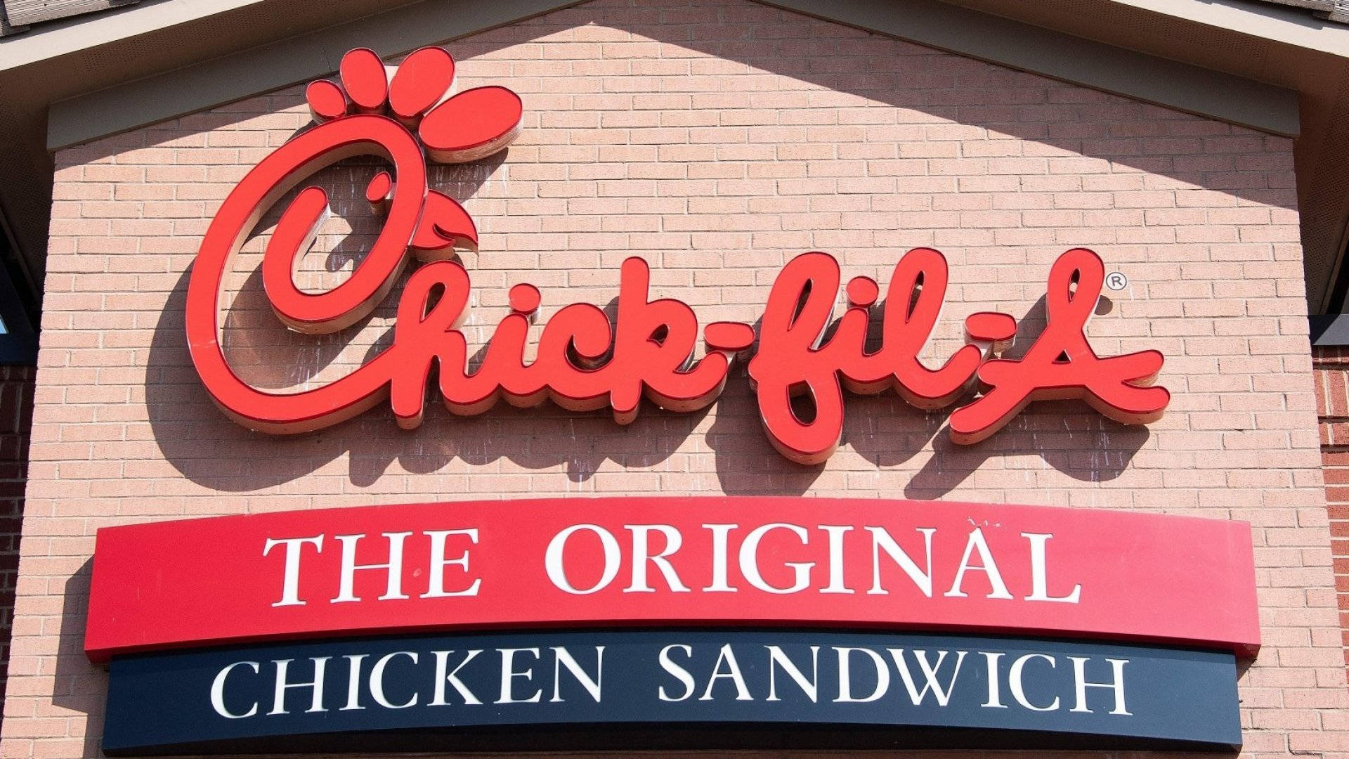 Chick Fil A 經典招牌 壁紙 — 一個鮮紅色的Chick-fil-A標誌，帶有粗體字，安裝在磚砌建築上，在晴朗的日光下。