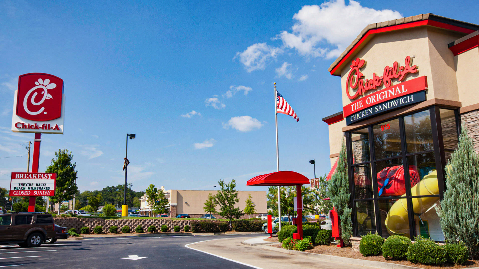 Chick Fil A 駕駛取餐區 壁紙 — 晴朗藍天、蓬鬆白雲、飄揚美國國旗下明亮的Chick-fil-A餐廳。