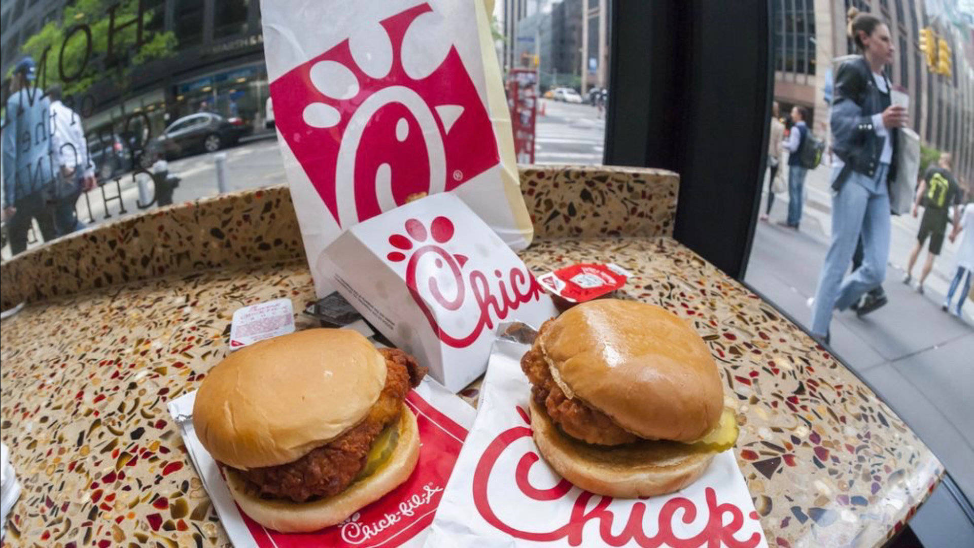 Chick Fil A 魚眼鏡頭拍攝 壁紙 — 兩塊香脆雞肉三文治放在有Chick-fil-A標誌的斑點桌面上，從繁忙的城市餐廳內觀看。
