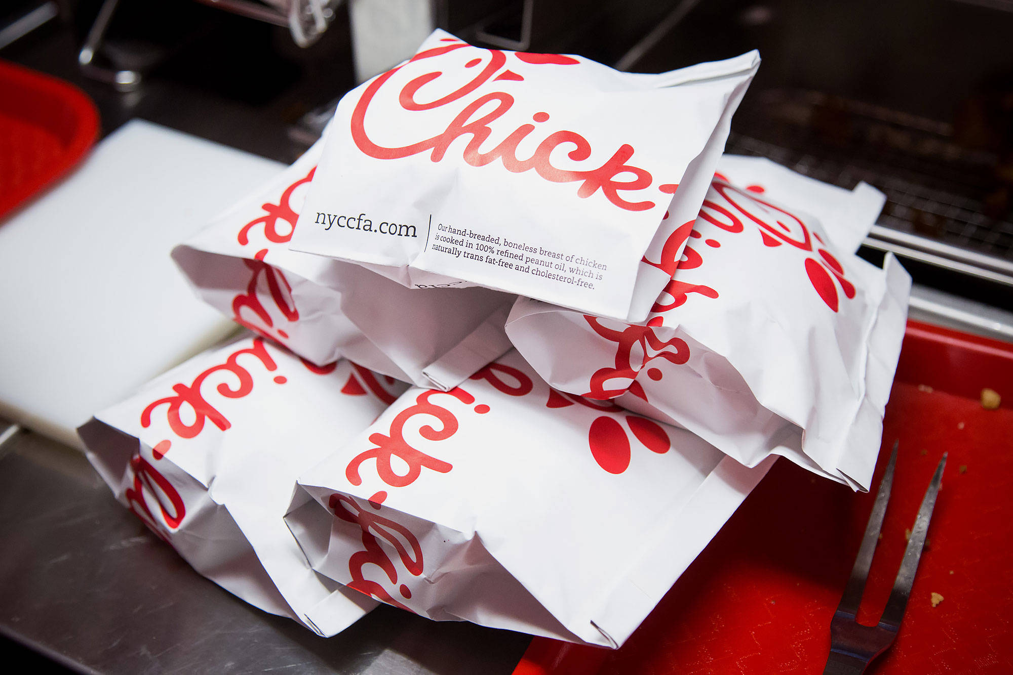Chick Fil A外帶餐點 壁紙 — 一堆帶有紅色標誌的白色Chick-fil-A紙袋放在快餐廚房的金屬叉旁邊的紅色托盤上。