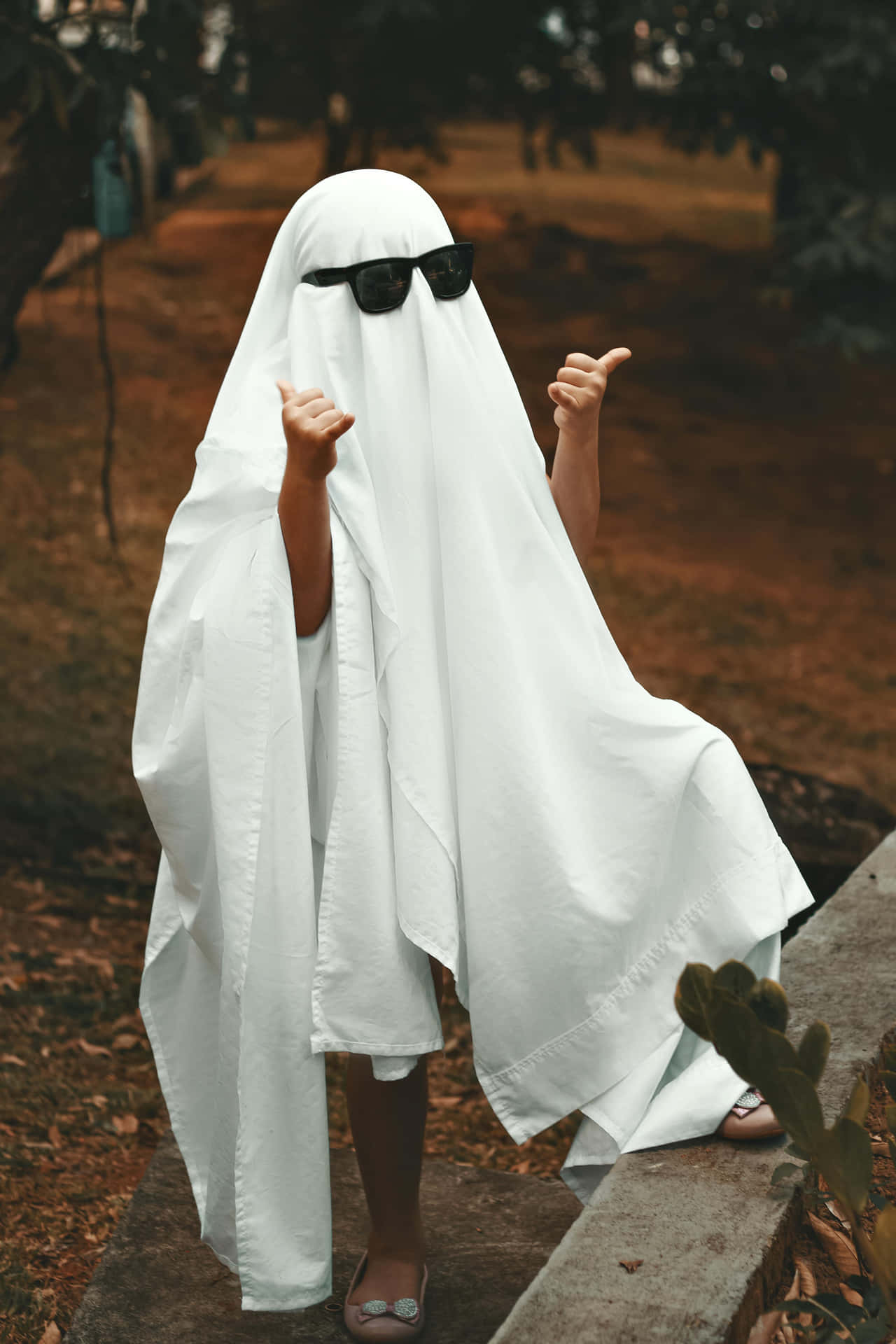  壁紙 — 一個穿著白色鬼魂服裝、戴著太陽眼鏡的小孩，站在戶外的石頭邊緣上，豎起兩個大拇指。
