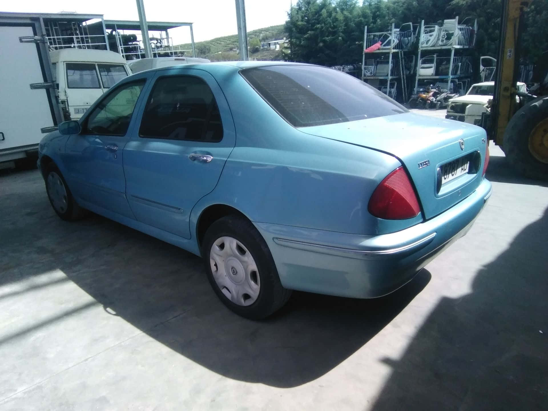  壁紙 — 一架淺藍色房車停泊在陽光普照的戶外場地，背景有工業設備和樹木。