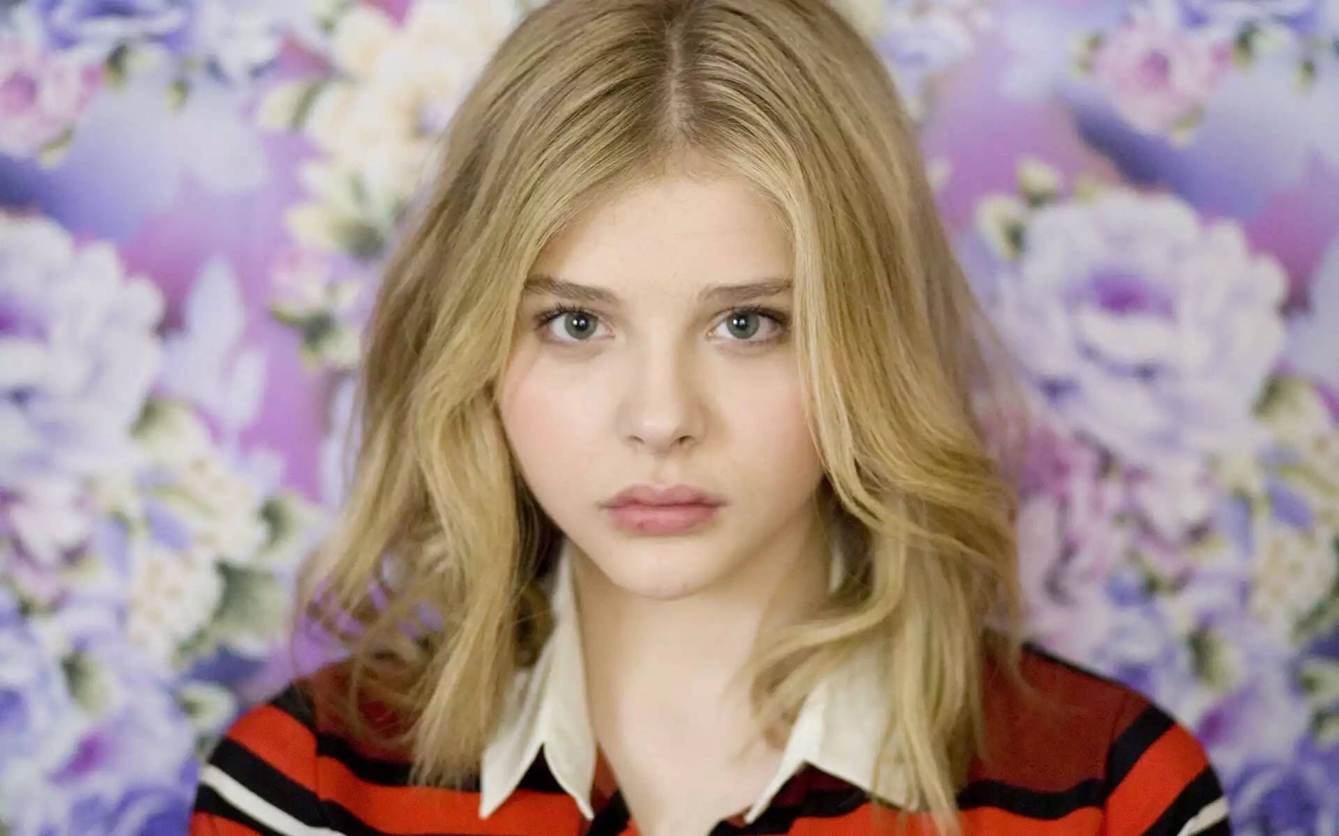 凝視的 Chloë Grace Moretz 壁紙 — 一位金髮藍眼的年輕女子在充滿活力的紫色花卉壁紙背景下直視鏡頭。