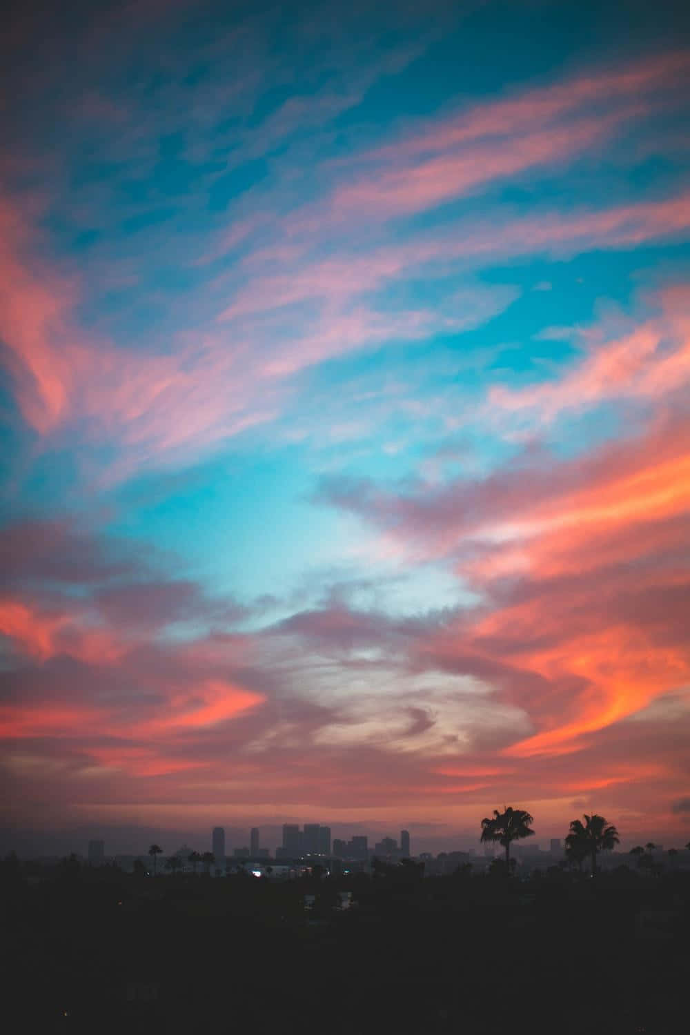 Colorful Sunset 桌布 壁紙 — 充滿活力的日落天空，粉紅色、橙色和藍色的雲朵勾勒出城市天際線和棕櫚樹的剪影。