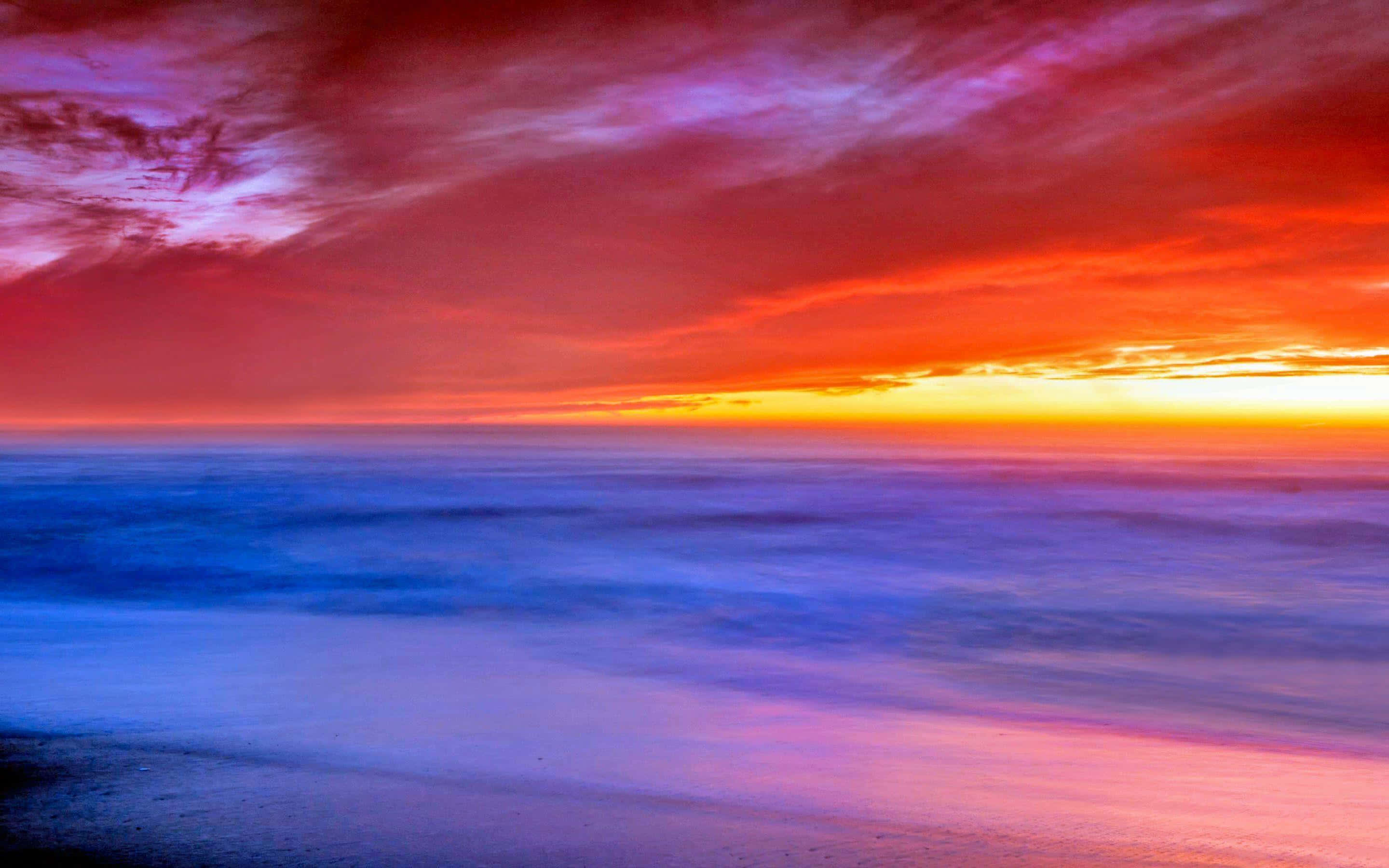 Sunset Landscape 桌布 壁紙 — 充滿活力的日落將天空染成火紅和橙色，映照在平靜的海洋上，伴隨著模糊的藍色波浪。